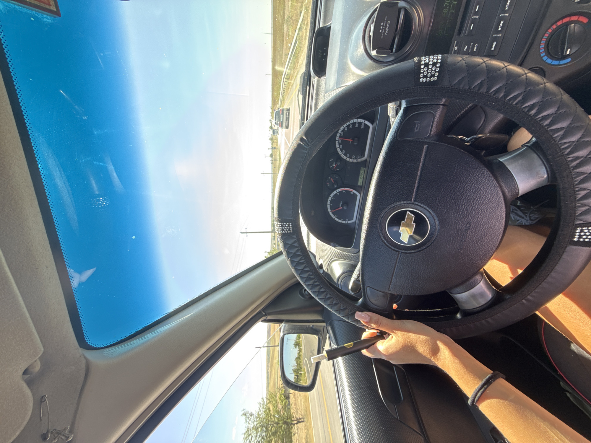 A person's hand holds a black and gold electronic cigarette in the driver's seat of a car. The car is moving on a road, indicated by the blurred background and the perspective of the view through the windshield. The dashboard and steering wheel are visible. The steering wheel has a black cover with diamond-like embellishments and a Chevrolet logo in the center. The sun is shining brightly, casting shadows on the dashboard and the driver's hand. The exterior view through the windshield shows a road lined with vegetation and utility poles, with other cars in the distance. The weather appears to be clear and sunny. The location is identified as Cioc-Maidan, Moldova, suggesting a rural or semi-rural setting.