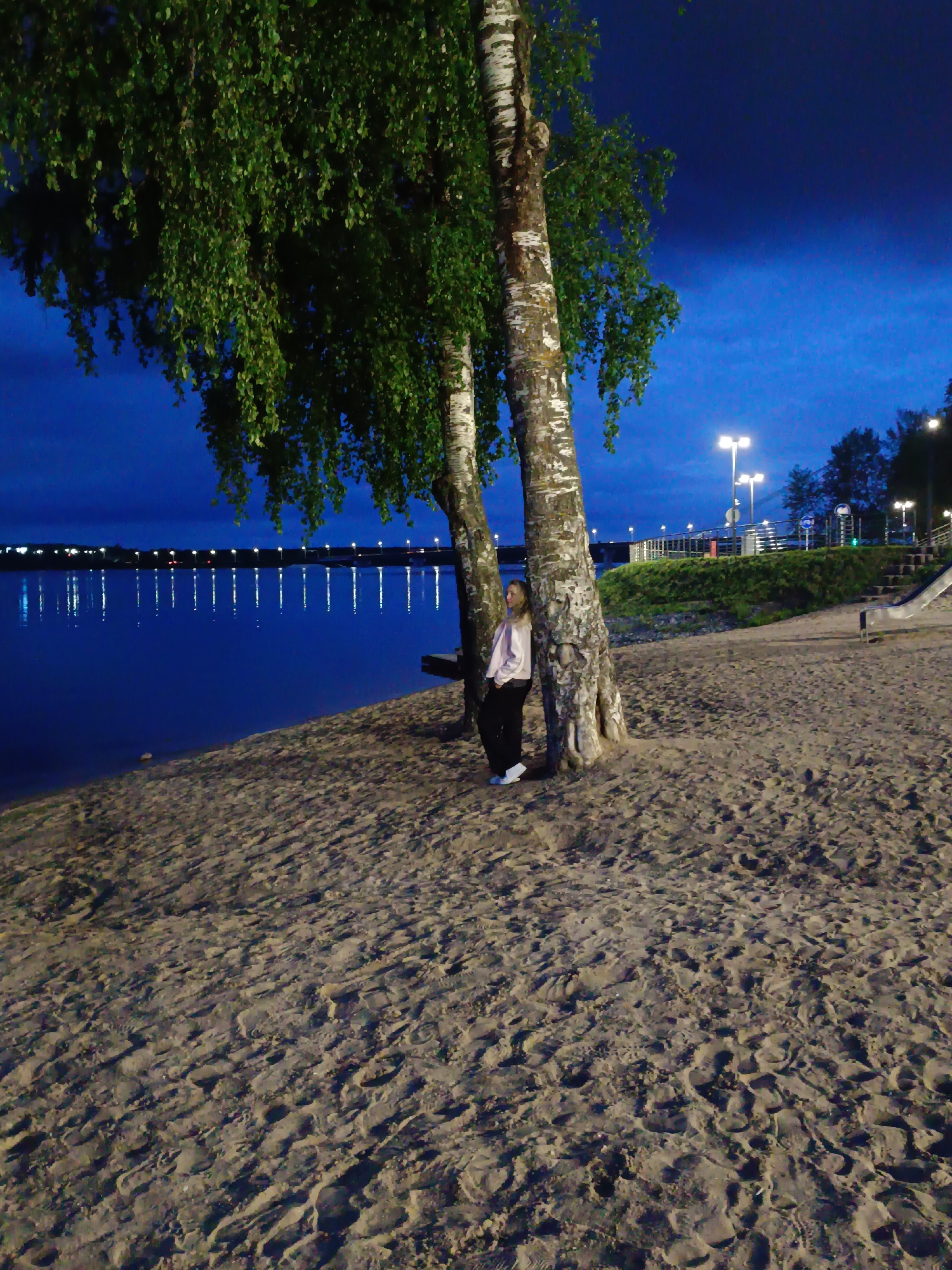 The image captures a tranquil night scene along a sandy beach beside a wide body of water, likely a river or lake. In the foreground, two prominent birch trees with characteristic white bark and dense green foliage stand near the water's edge.
A woman is the central human subject, leaning casually against one of the birch tree trunks. She has light-colored hair and is dressed in a light-colored long-sleeved top, dark trousers, and white sneakers. Her gaze is directed towards the left, across the water.
The sandy beach, stretching from the bottom of the frame to the waterline, is marked with numerous footprints. The water, a deep blue, reflects a long line of bright, distant lights from what appears to be a bridge or embankment across the expanse. On the right side of the frame, beyond the beach, a grassy bank rises, featuring streetlights and the visible structure of a metal slide, suggesting a recreational area or park.
The sky is a dark, clear blue, indicating either deep twilight or nighttime. The artificial lights from the distant bridge and the streetlights illuminate parts of the scene, creating bright reflections on the calm water. The overall atmosphere is quiet and contemplative. No visible text is present in the image.