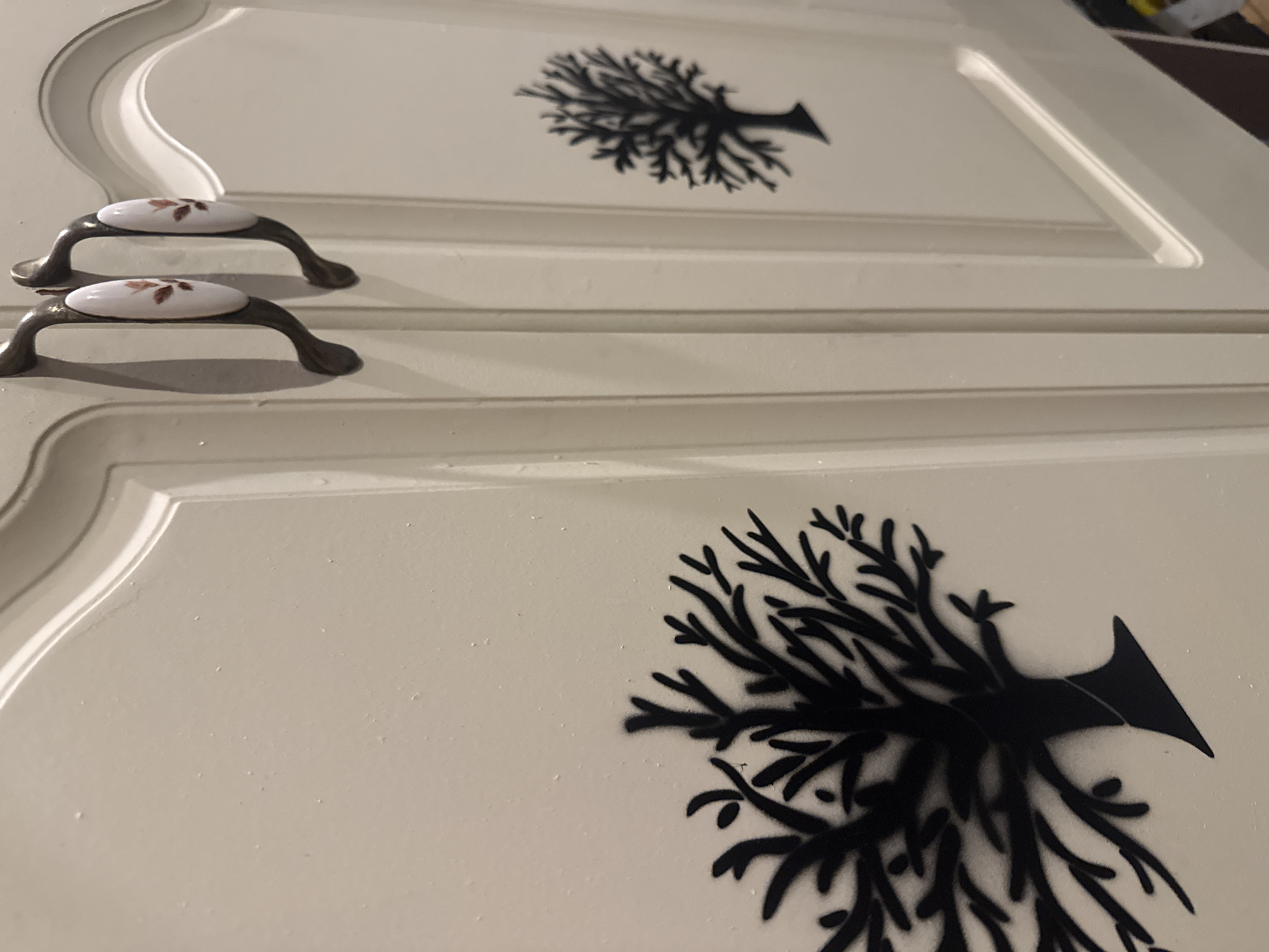 A close-up, eye-level shot of two cream-colored cabinet doors reveals decorative elements. The door on the left features a large, black stencil of a stylized tree with intricate branches filling a significant portion of its surface. The door on the right, positioned slightly further back and to the right, displays a smaller, identical black stencil of a tree. Both doors have ornate, dark metal handles with oval, white ceramic inserts, each decorated with a delicate pattern of small, reddish-brown leaves. The cabinet doors appear to be part of a larger piece of furniture, possibly in a home environment, with subtle highlights and shadows indicating indoor lighting. The overall aesthetic is one of rustic charm and understated elegance. There is no indication of the specific location within Galinė, Lithuania.
