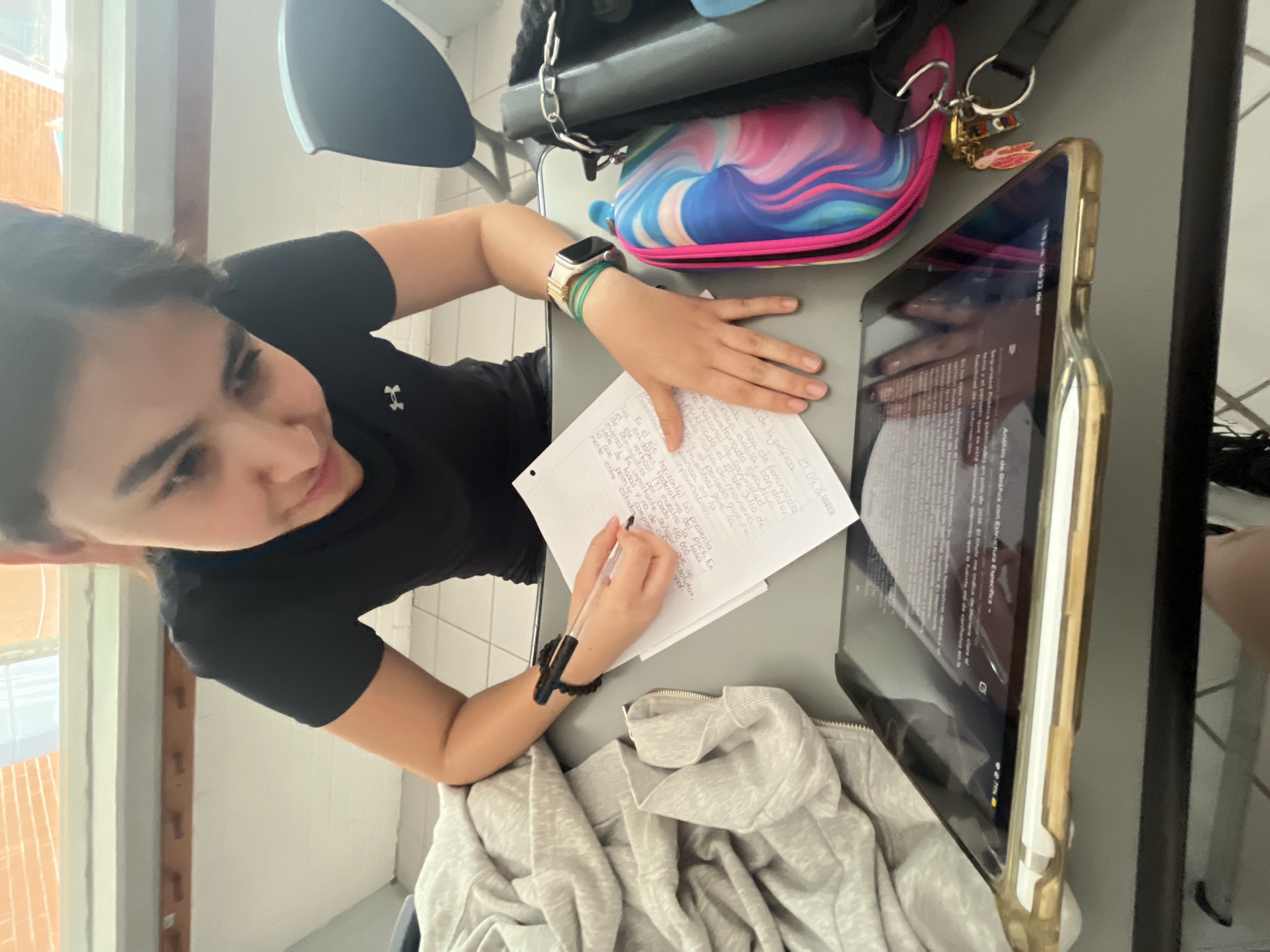 A young woman sits at a desk, writing on a piece of paper. To her right is a tablet displaying text, while other items such as a purse, a pencil case, and a sweater are also present on the desk. She is wearing a watch on her left wrist and a bracelet on her right. The location is a room with a window.