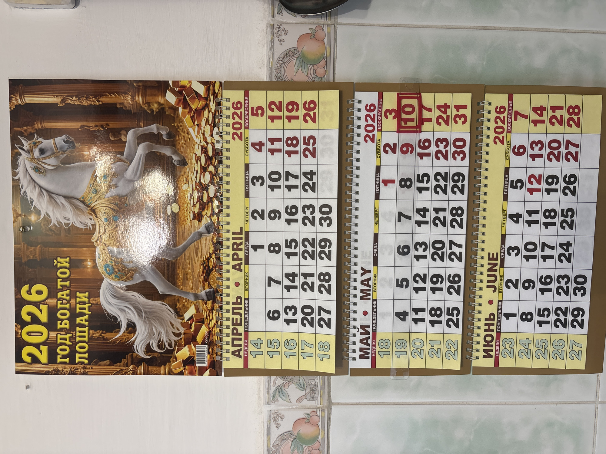The image shows a 2026 calendar, featuring a carousel horse image on the cover. The open pages display the months of April, May, and June 2026. May 10th is circled in red on the May page. The calendar is resting on a tiled surface with decorative tile accents. There is no evidence to suggest it is in Zagatuy, Russia.