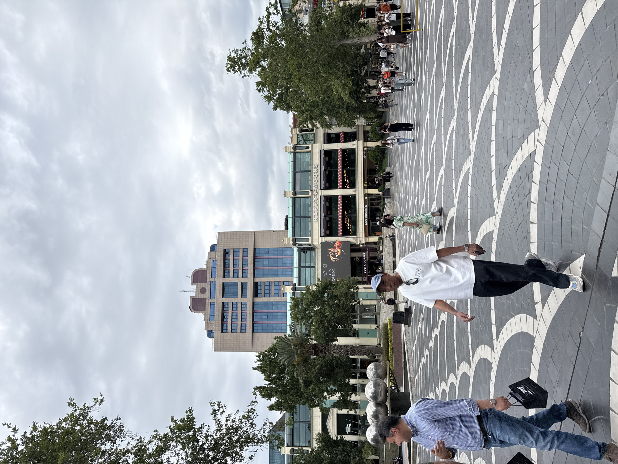 This media file captures a lively outdoor scene with several people visible. In the foreground, two men are walking. One, wearing a blue checkered shirt and jeans, is carrying a black shopping bag. The other, in a white t-shirt and dark pants, has sunglasses on the back of his head. Behind them, a patterned paved walkway leads towards a modern building complex. This complex features storefronts, including a McDonald's and a Costa Coffee. Trees and palm trees add greenery to the urban setting. The sky is overcast, suggesting a cloudy day, and the lighting indicates it is daytime. The overall atmosphere appears casual and public, as if people are going about their daily activities or enjoying a stroll. The architectural style of the building and the paving suggest a contemporary urban environment.