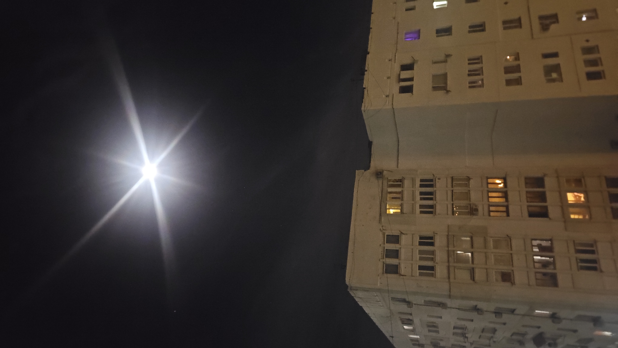The image presents a striking nighttime view of an urban environment, likely in Sevastopol, Ukraine, captured from a low, upward-looking perspective.
A dominant and intensely bright light source, possibly the moon or a powerful streetlamp, is positioned in the upper-left quadrant of the frame. It emits a brilliant white light with a pronounced starburst effect, casting long, sharp rays across the dark, clear night sky. The sky itself is a deep, uniform black, indicating the late hours.
To the right, a large, multi-story building occupies the other half of the image. The building appears to be an apartment block or a large institutional structure, characterized by its light-colored facade, possibly concrete or stucco, and a utilitarian architectural style that might be typical of post-Soviet urban developments. The building's structure is complex, appearing to have multiple sections or wings with various setbacks and projections. Numerous rectangular windows are visible across its many floors. Many of these windows are illuminated from within, displaying a mix of warm yellow, cool white, and notably, one window with a distinct purple glow, suggesting ongoing activity or habitation.
No people or explicit activities are visible, and there is no discernible text in the frame. The image conveys a serene yet imposing nocturnal scene, highlighting the contrast between the artificial light from the building and the singular, powerful natural or artificial light source against the dark canvas of the night sky.