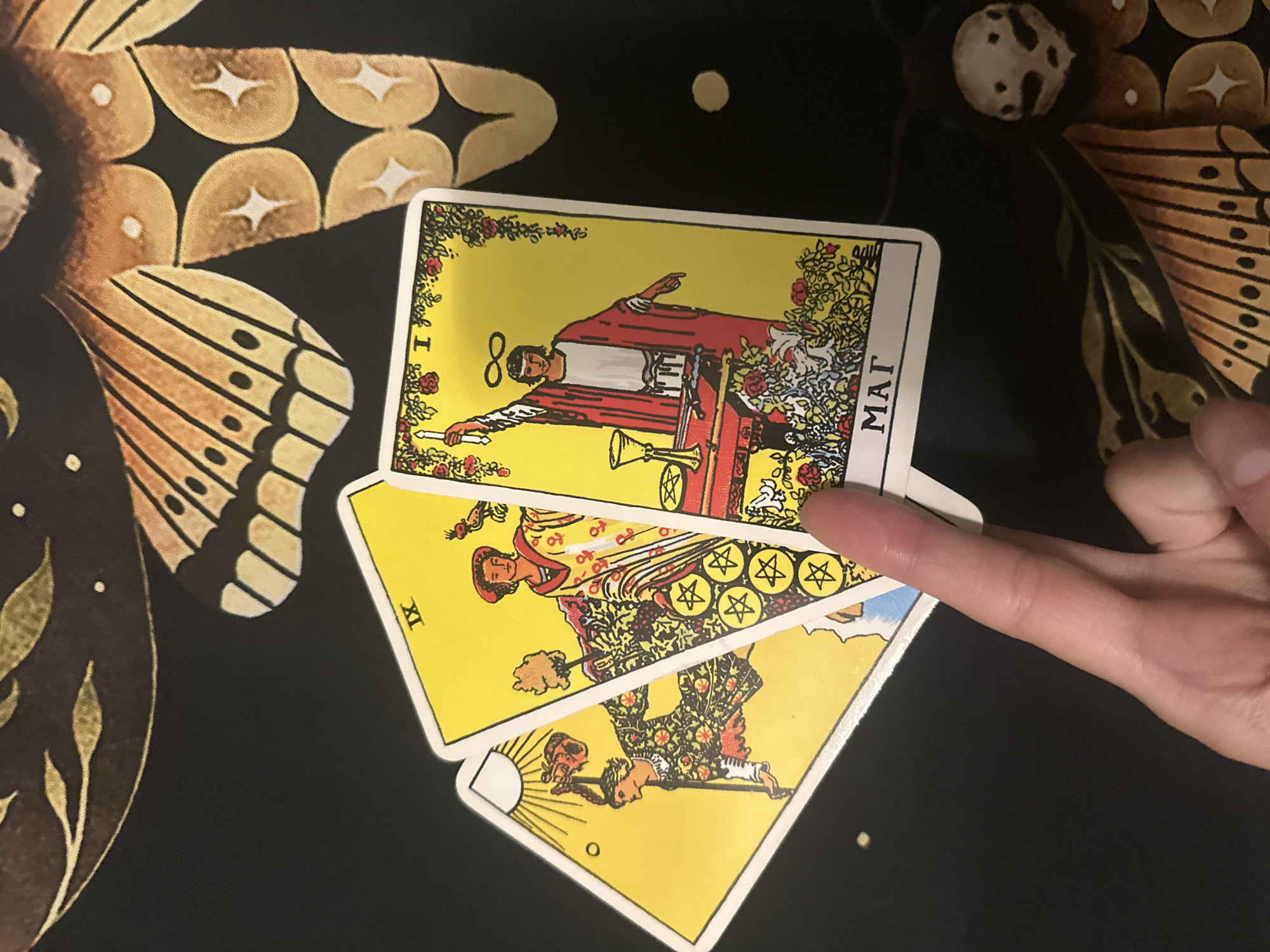 The image shows a close-up of three tarot cards laid out on a black cloth with a butterfly wing pattern. A person's finger is holding the cards, obscuring the bottom right portion of the card on the right. The cards visible are:
1. **The Fool (numbered 0):** Depicts a young person with a bag on a stick, stepping towards a cliff under a bright sun. This card is on the far left and partially obscured.
2. **Nine of Pentacles (numbered IX):** Shows a woman in a rich robe standing in a vineyard, holding a falcon and surrounded by pentacles. This card is in the middle.
3. **The Magician (numbered I):** Features a figure with a staff raised, an infinity symbol above their head, and symbols of the four suits on a table before them. This card is on the right.
The text "MAГ" is visible at the bottom of The Magician card. The background cloth is black with an abstract, golden-yellow and brown pattern resembling butterfly wings. There are no discernible elements to suggest a location in Togliatti, Russia.