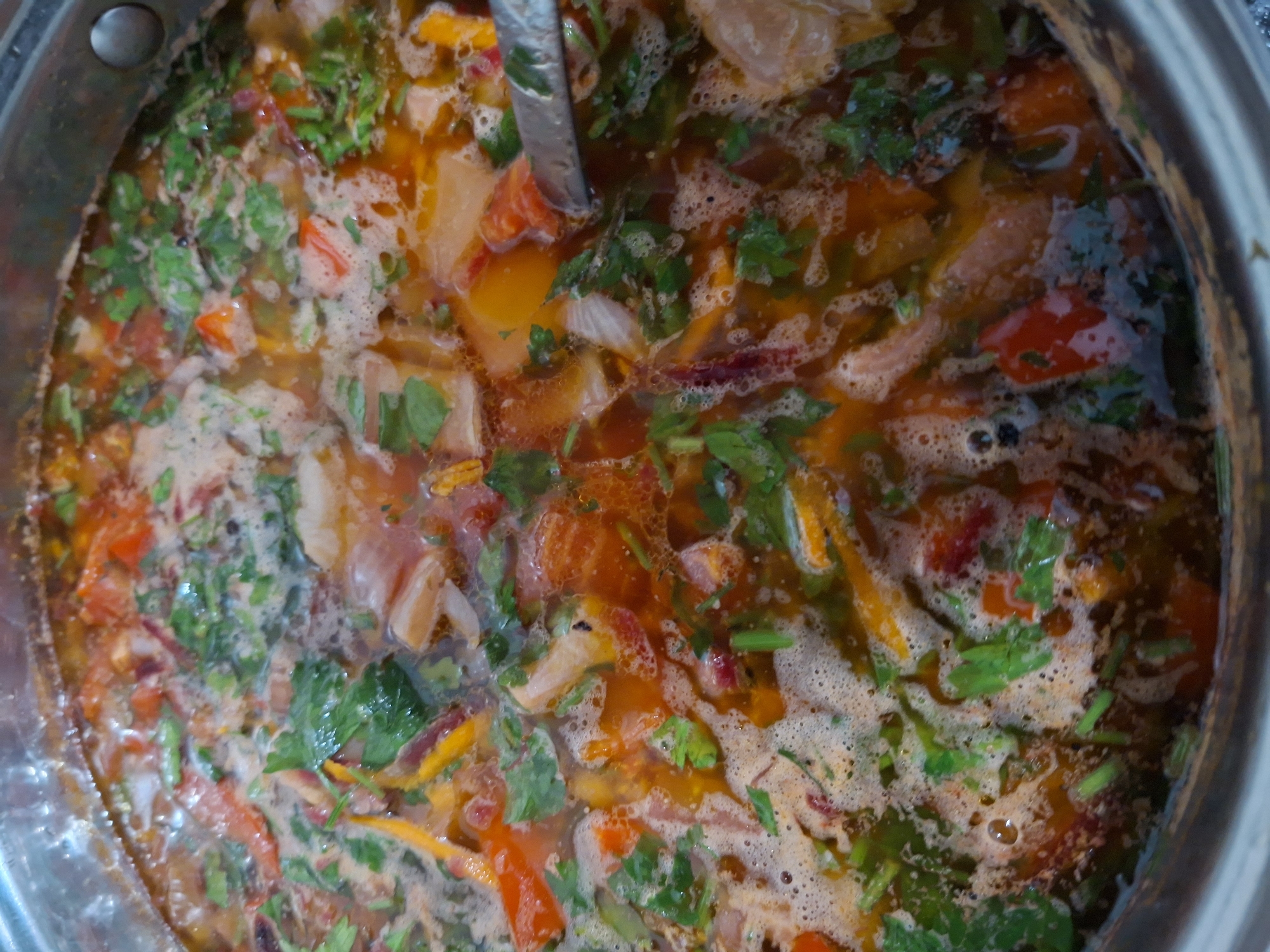 The image shows a close-up view of a pot of soup, likely a hearty stew or broth, filled with various ingredients and bubbling on a stovetop. The soup is a rich, orange-red color, suggesting the presence of tomatoes, carrots, or other vegetables. Visible ingredients include chopped tomatoes, pieces of what appear to be meat or poultry, and fresh green herbs, possibly cilantro or parsley, scattered throughout. Thinly sliced onions are also discernible. The soup is actively cooking, with a layer of foam on the surface and small bubbles indicating heat. A metal ladle or spoon is partially submerged in the soup on the left side of the frame, suggesting someone is in the process of stirring or serving. The pot itself is metallic and appears to be made of stainless steel, with a visible rim. The lighting suggests an indoor setting, likely a kitchen, with the focus entirely on the food being prepared. The overall impression is one of a home-cooked meal being made, possibly in Chișinău, Moldova, given the location context. There is no visible text in the image.