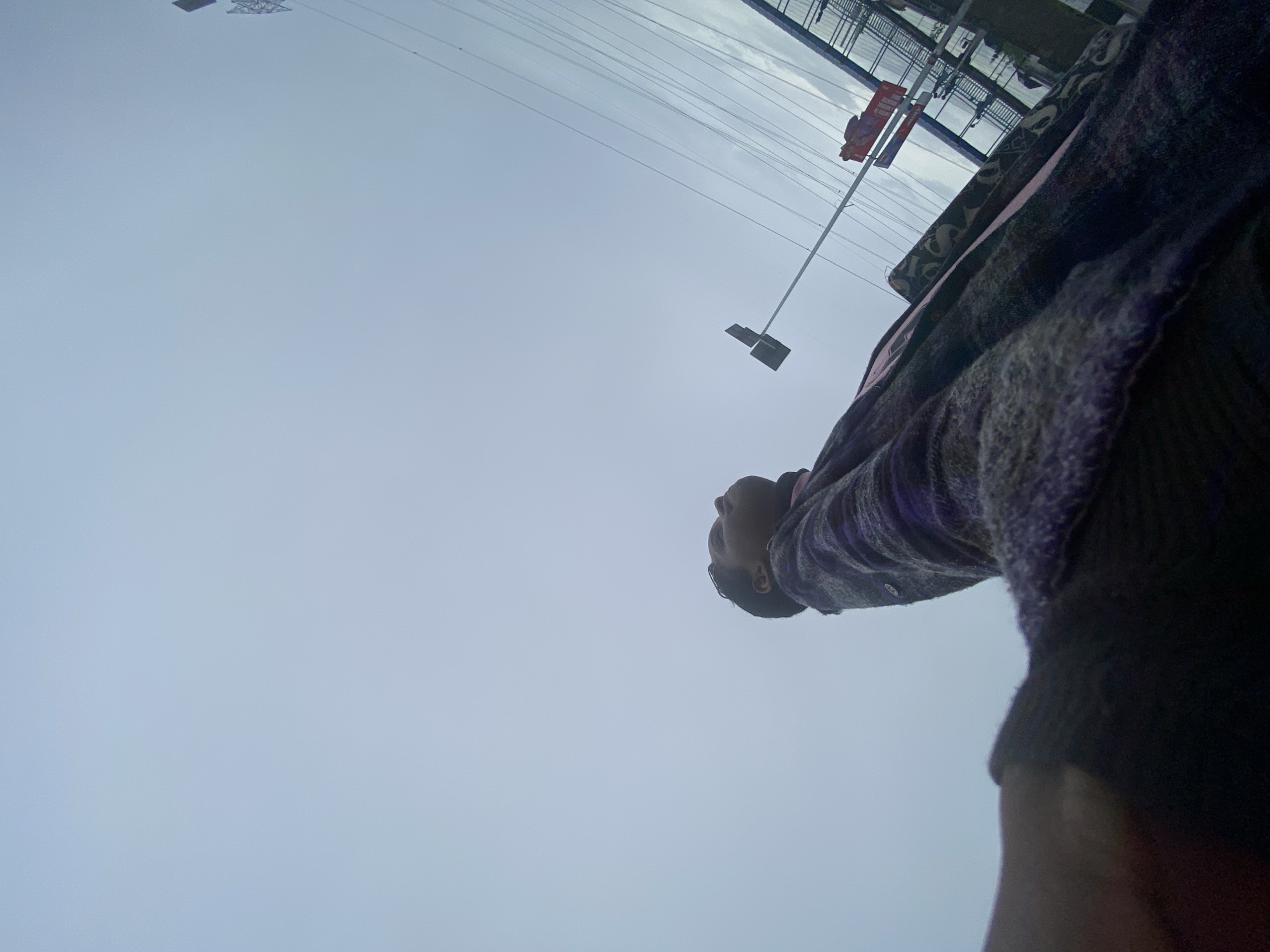 A low-angle shot looking upwards at a person standing outdoors in what appears to be an urban environment. The person is wearing a dark-colored sweater or jacket with a patterned design. Above the person, there is a cloudy sky and a glimpse of a building or structure with exposed metal framework and power lines. In the upper part of the frame, a red banner or sign is visible.