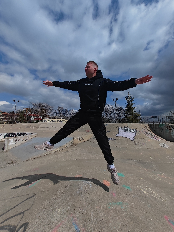 A male individual is captured mid-air in a skatepark in Shumen, Bulgaria, against a partially cloudy sky. He is positioned centrally, leaping with his arms extended horizontally and legs spread wide apart, forming an "X" shape. The individual wears a black hooded sweatshirt with white piping along the sleeves and hood, and black cargo pants. A white logo, possibly "OUTERSTVND," is visible on the front of his hoodie. His feet are clad in light beige or grey athletic shoes with white socks. His head is turned to the right, and he appears to be looking in that direction. A distinct, elongated shadow of his airborne form is cast on the grey concrete surface of the skatepark below him.
The skatepark environment features various concrete ramps, ledges, and transitions, all covered in graffiti. Visible graffiti includes text such as "HOMAY," "AYOUI," "GDS," "STAY," "NICKAS," and "COOL," alongside abstract shapes and a cartoon-like creature resembling a white, blob-shaped figure with black eyes and a mouth. Patches of colored chalk or paint are also present on the ground. In the background, deciduous trees, some with visible bare branches, are present. Further back, residential buildings with terracotta-colored roofs and tall street light poles are discernible. The sky above is characterized by a mix of bright white and darker grey cumulus clouds, indicating a partially sunny day.