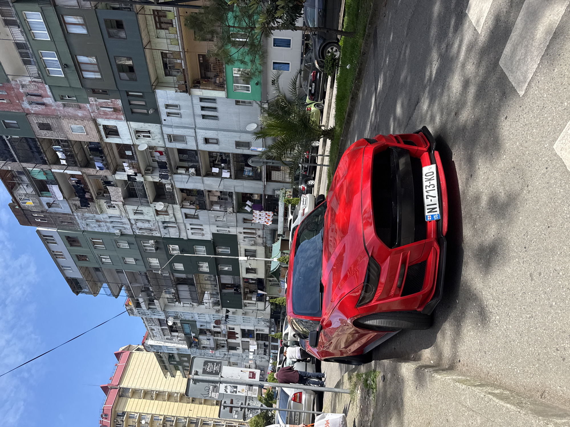 The image shows a red sports car parked on a street in front of a tall, weathered apartment building. The building appears to be in a densely populated urban area. The car is facing away from the camera, and its license plate, "NI-713-KO," is visible. Other cars are parked along the street. There are pedestrians visible. The image suggests an urban environment, likely a city, with older architecture.