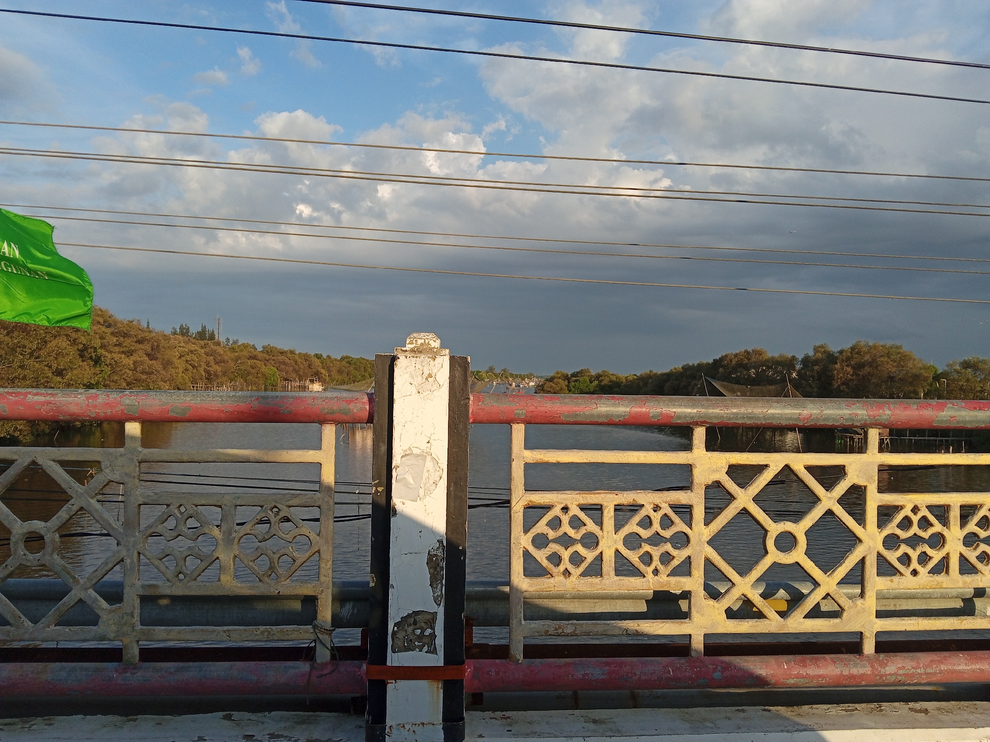The image depicts a bridge railing in Lhokseumawe, Indonesia, overlooking a body of water. The railing is constructed of weathered concrete or painted metal with a decorative geometric pattern. The paint is peeling and shows signs of age and wear. A central pillar is partly shadowed. Beyond the railing, there is a view of the water, the opposite bank with trees and vegetation, and a sky with clouds. Power lines run across the top of the frame. To the left, a green flag is visible.