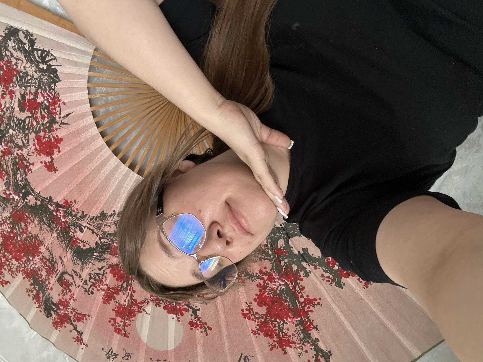 The image features a person, likely female, lying down indoors, in Olkhovka, Russia. She has long brown hair and is wearing a black t-shirt. Her face is partially obscured by large, fashionable glasses with highly reflective blue lenses. One hand, showing white-tipped fingernails, rests near her head. Her expression appears calm and relaxed, with a subtle hint of a smile.
A prominent, large, open folding fan is positioned underneath or beside her. The fan displays a delicate pink background patterned with vibrant red cherry blossoms and dark, intricate branches, suggestive of East Asian art. The wooden ribs of the fan are visible on the left side. Faint, stylized black calligraphy, resembling Asian characters, is visible on the pink surface of the fan near the bottom right. The person is posing for a photograph, creating a relaxed portrait shot. The setting appears to be an indoor environment with a light-colored, possibly textured, flat surface in the background.