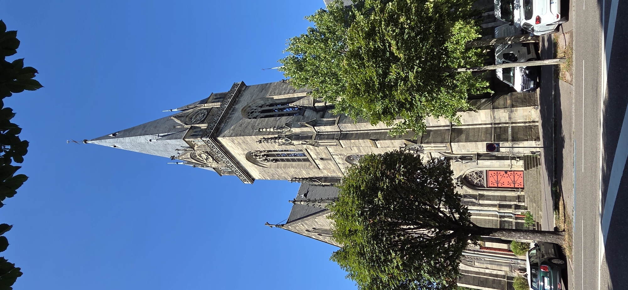 The image captures a daytime view of a stone church in Strasbourg, France. The church features a prominent tall spire with gothic architectural elements, including arched windows and decorative stonework. A clock face is visible on the tower. In front of the church, a red door with ornate detailing is set within an arched entryway, accessible by a set of stone stairs. Lush green trees partially obscure the lower sections of the church. Several vehicles are parked along the street in front of the building, including a white van and a white car. A dark-colored mini cooper is parked on the left side of the image, near a person standing by the church entrance. The sky is clear and blue, indicating good weather. The scene is framed by dark green leaves from trees in the foreground, suggesting the photo was taken from across a street or sidewalk.