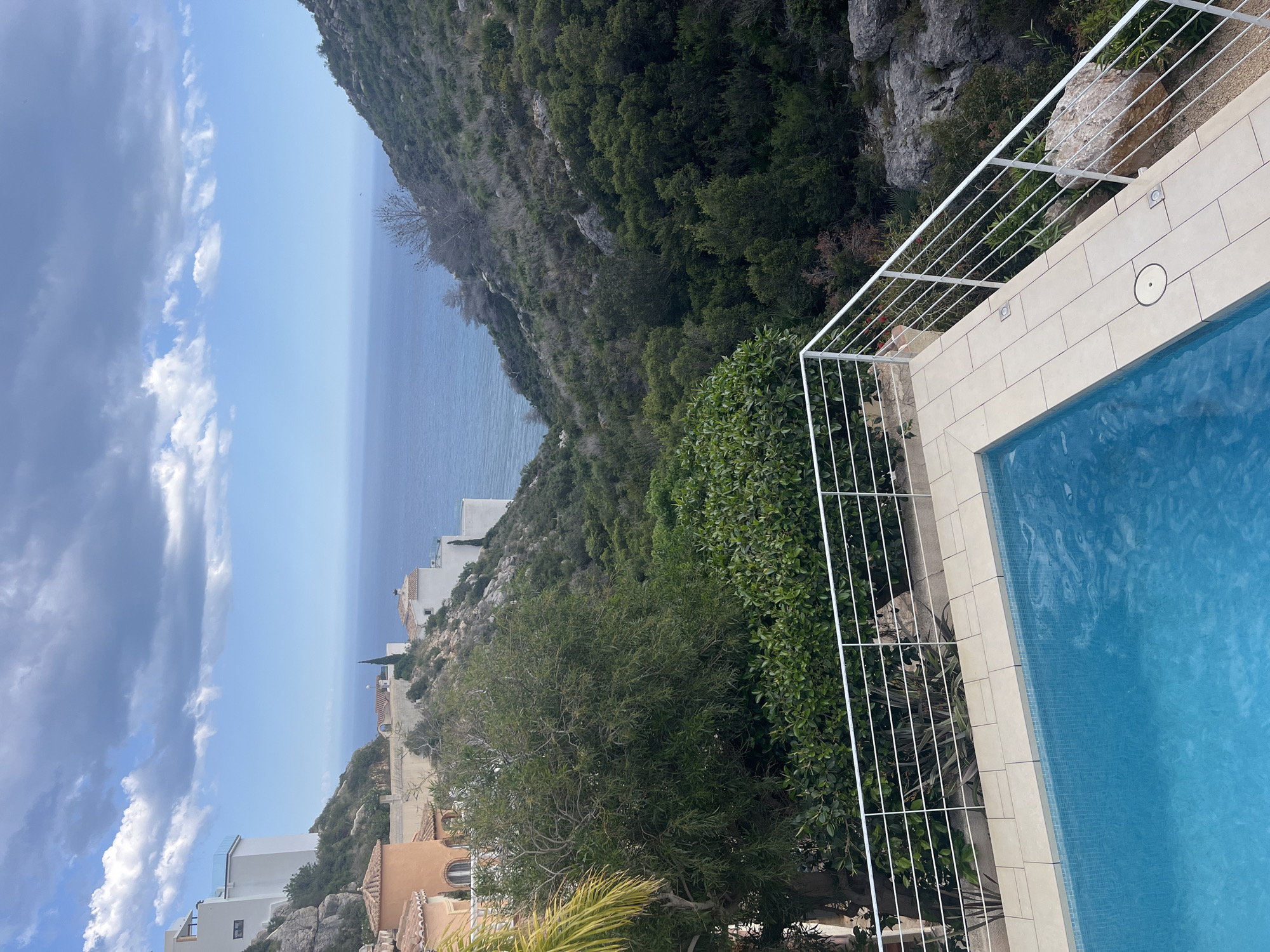 The image shows a view from a residential area overlooking the Mediterranean Sea, most likely in the municipality of El Poble Nou de Benitatxell / Benitachell, Spain, based on the coastal hillside architecture and vegetation. In the foreground, part of a swimming pool with light blue water is visible, enclosed by a white metal railing. Tiled paving stones surround the pool. Beyond the railing, the hillside is densely covered with green trees and shrubs. Houses and other buildings are visible on the slope leading down towards the sea. The sea stretches to the horizon under a bright blue sky with some scattered clouds.