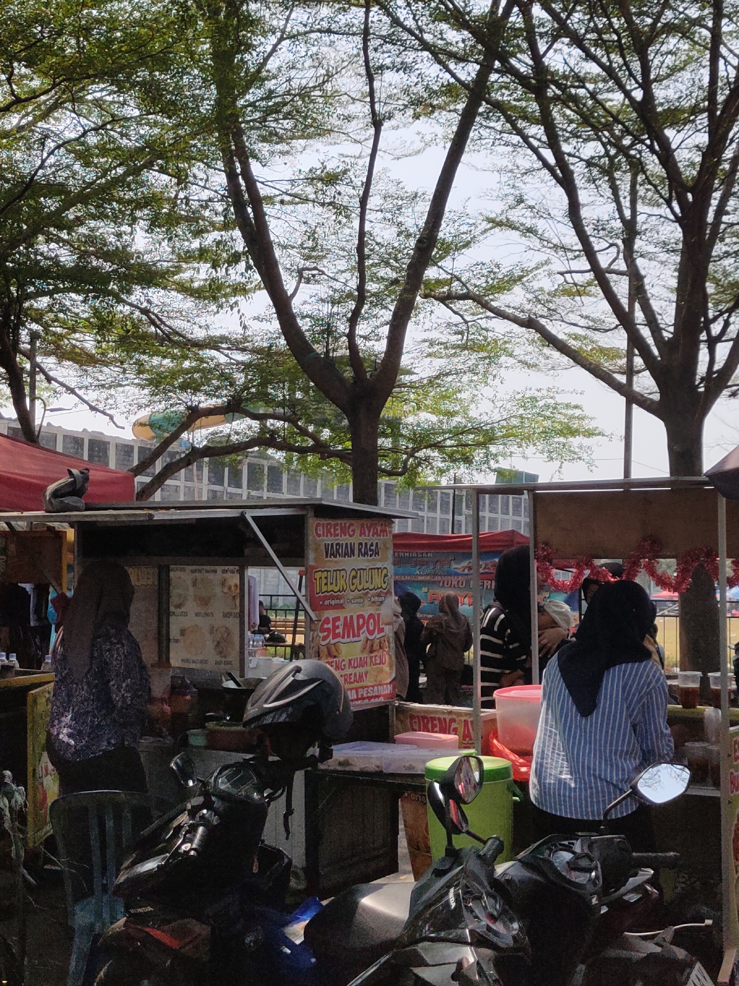 The image depicts a street scene in Ngasem, Indonesia, characterized by a row of food stalls under trees. In the foreground, several motorcycles are parked. One motorcycle in the center foreground is prominent, with a black helmet placed on its handlebars. Behind the motorcycles, food stalls are set up. One stall displays a sign for "CIRENG AYAM VARIAN RASA TELUR GULUNG" and "SEMPOI." Several individuals are visible, including a person in a hijab and a striped shirt standing behind a stall, and another person wearing a hijab and a patterned top to the left. A cat is perched on the roof of a stall on the left side of the image. In the background, more stalls and trees are visible, with a partial view of a building and a yellow and orange structure that appears to be a slide or a play structure. The overall impression is of a bustling local market or food vendor area.