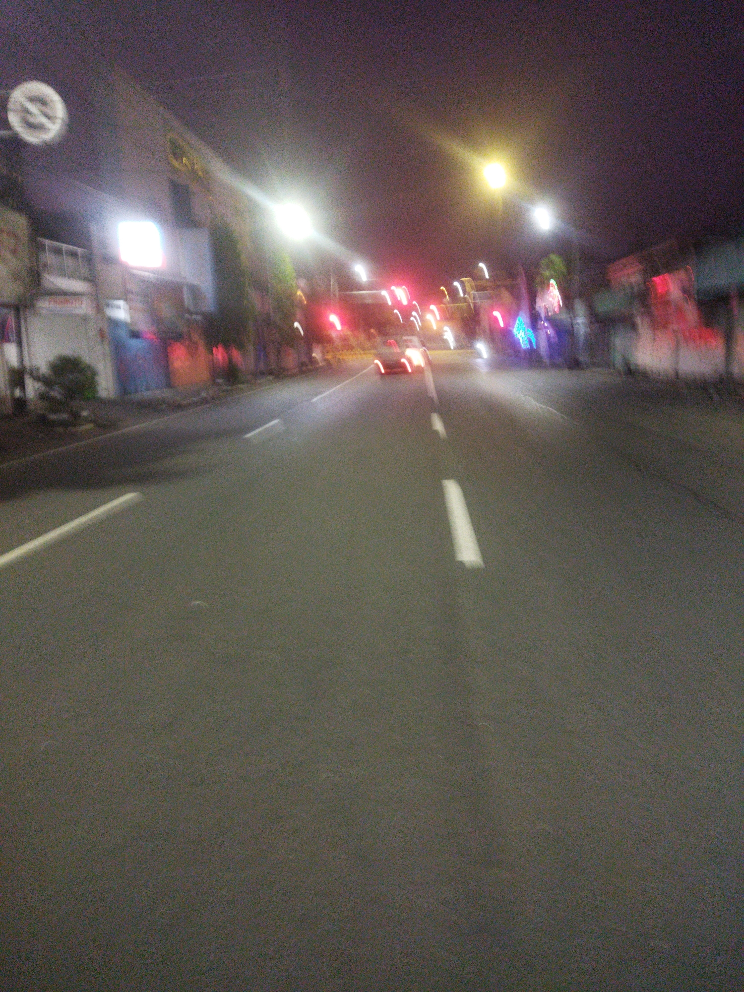 This is a night-time, blurred image of a street in Giripurwo, Indonesia. The perspective is from a moving vehicle, looking forward down a road with asphalt pavement. White dashed lines mark the lanes on the road. On the left side of the road, there are buildings with illuminated signs and some trees. On the right side of the road, more buildings and trees are visible, also with lights. There are several vehicles in motion on the road, indicated by blurred lights. The sky is dark and appears to be overcast. A circular sign with a diagonal line through it is visible on a pole on the left side of the frame. The overall impression is of a busy street at night with artificial lighting.