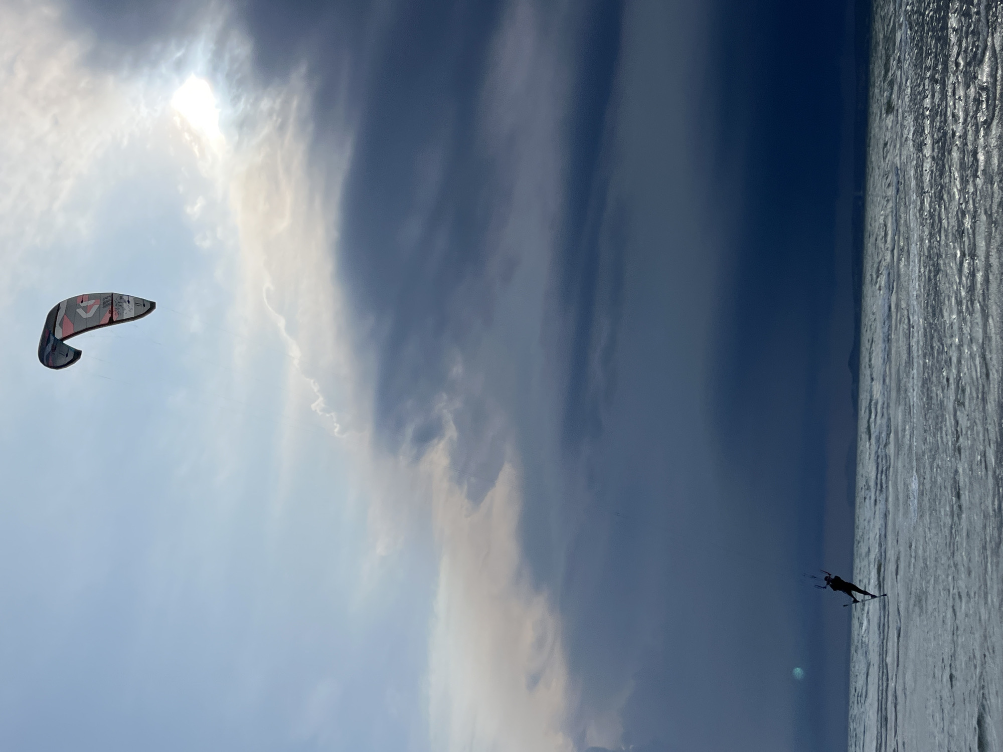 This media file captures a dynamic scene of kitesurfing in Mũi Né, Vietnam. The image features two main subjects: a colorful kitesurfer and a kitesurfing kite. The kitesurfer, seen as a silhouette against the water, is actively engaged in the sport, appearing to be in mid-action. The kite, a large and predominantly gray and red object, is soaring in the sky to the left of the frame, suggesting it is providing the power for the surfer.
The setting is a coastal area, with the choppy, textured surface of the ocean occupying the right side of the image and a vast sky filling the rest. The sky is a mix of dramatic clouds, with patches of bright sunlight breaking through, indicating a time of day with strong illumination, likely midday or afternoon. The weather appears to be windy, as evidenced by the kitesurfing activity and the cloud formations. The presence of the ocean and the activity of kitesurfing strongly point to a beach or a coastline known for watersports.
Notable details include the way the sun is partially obscured by clouds, creating a luminous effect in the upper left quadrant. The water's surface is rendered with a rough texture, reflecting the light and emphasizing the windy conditions. The kitesurfer's posture suggests an energetic and possibly exhilarating moment.
There is visible text on the kite, though it is partially obscured and difficult to read clearly due to the image resolution and angle. Some lettering and logos are discernible, hinting at a brand or design. The overall mood conveyed is one of adventure and the power of nature.