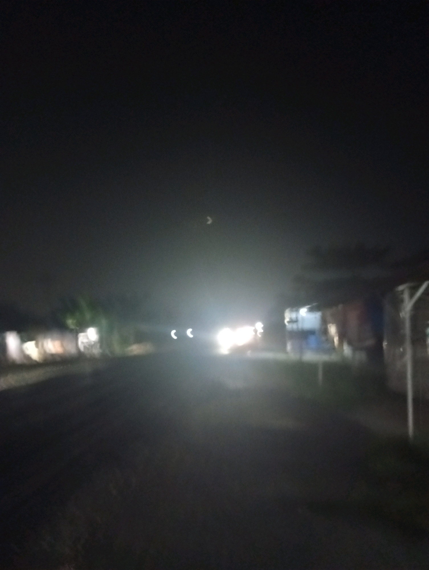 The image captures a dimly lit road at night, possibly in Kubang Jaya, Indonesia. The scene is characterized by poor visibility due to darkness and what appears to be fog or dust in the air, creating a hazy atmosphere. In the distance, the headlights of a vehicle are visible, casting a bright glare and two distinct circular lights that suggest the vehicle is approaching. On either side of the road, indistinct shapes of buildings or structures can be seen, with some exhibiting faint interior lights. A crescent moon is faintly visible in the upper part of the sky. The road surface itself is dark and appears to be unpaved. The overall impression is one of a quiet, rural road at night, with limited activity and the presence of a single approaching vehicle.