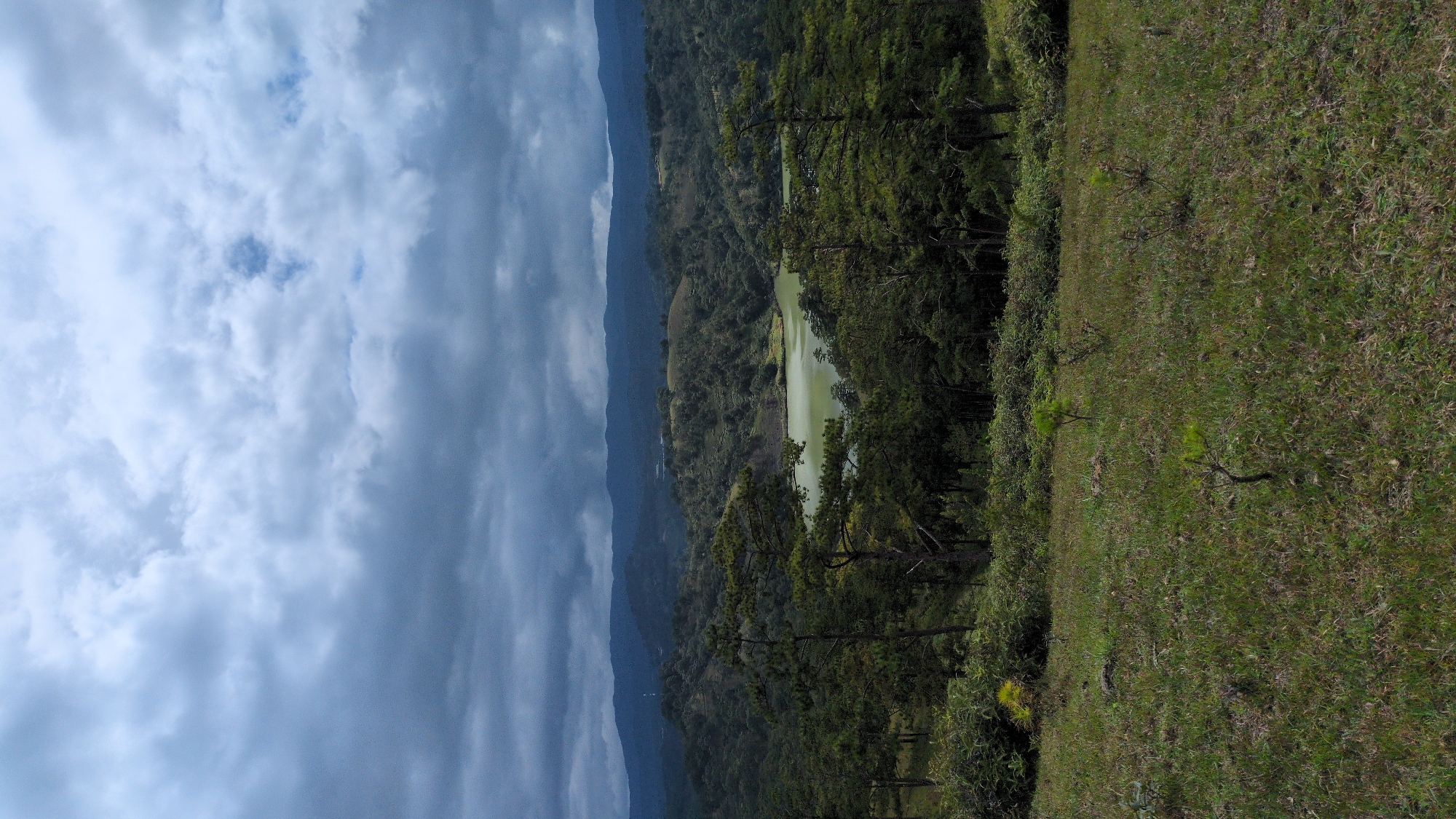 A vertical, wide-angle shot captures a scenic view of a lake nestled amongst rolling hills covered in dense forest, under a dramatic, cloudy sky. The foreground features a grassy slope with scattered small trees and patches of dry grass. In the mid-ground, a light green lake is visible, surrounded by a mix of pine and other trees. The background consists of layered, dark green hills and mountains under a heavy, overcast sky with patches of lighter clouds. The location is identified as the City of Lang Biang, Da Lat, Vietnam, likely from an elevated viewpoint. There are no people or animals visible in the image. The overall atmosphere is serene and natural.