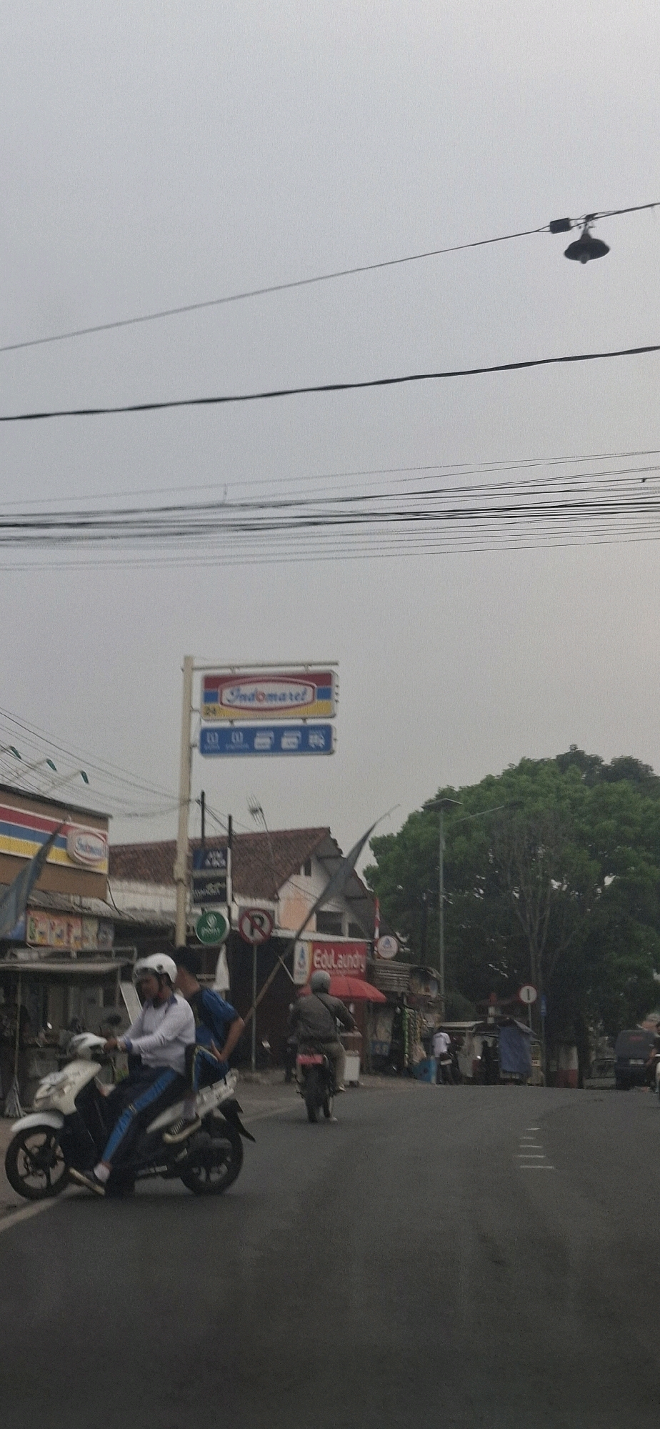 This is a vertical, eye-level shot of a street scene in Sirnagalih, Indonesia. The sky is overcast and gray, with multiple power lines crisscrossing the upper portion of the frame, and a hanging lamp fixture visible on the right. In the midground, a prominent red and blue sign for "Indomaret" is mounted on a pole, indicating a convenience store. To the left of the sign is a building with red tile roofing and a sign that says "Indomaret." Further left, a portion of another building with a similar red and yellow striped awning is visible. A white motorcycle with a rider wearing a white helmet and a white shirt is in the foreground, facing left and moving away from the camera. The rider is wearing blue pants. Another motorcycle with a rider wearing a brown jacket is further down the street, moving in the same direction. Several other buildings and structures line the street, some with awnings and makeshift stalls. A large, dense tree with dark green foliage stands to the right of the Indomaret sign, obscuring part of the background. The street is paved and appears to be a typical urban or suburban road with some traffic markings. The overall impression is a cloudy day in a busy commercial area.