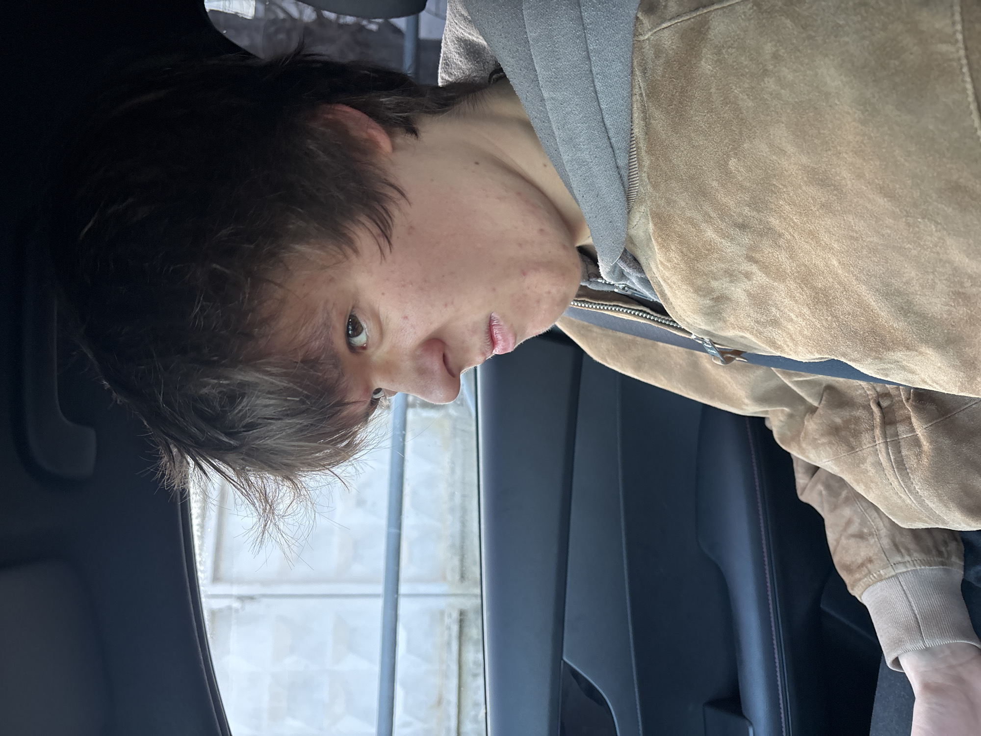 A person is shown in an indoor setting with part of their face visible. They have short, dark hair and appear to be wearing a brown jacket with a gray hooded sweatshirt underneath. The background shows a gray interior, possibly a car, with a window on the left side. The setting is unspecified, but the query mentions the city of Chaikovsky, Russia, without any of the objects indicating this is the correct location.