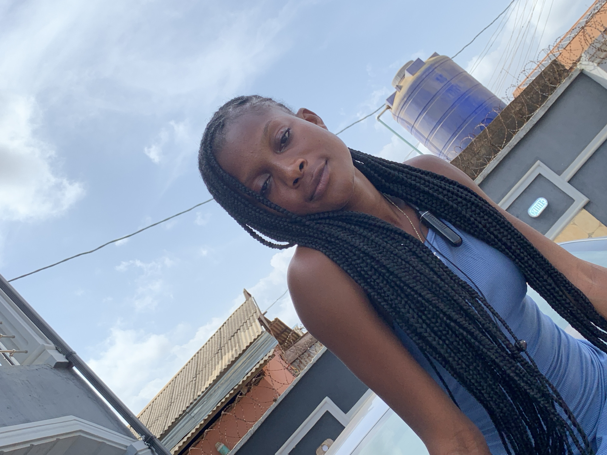 A low-angle shot shows a woman with braided hair wearing a blue dress. She has dark skin and a neutral expression. In the background are rooftops and buildings, including one with barbed wire. A large blue water tank sits atop a building. The sky is partly cloudy. Ikorodu, Nigeria is the location.