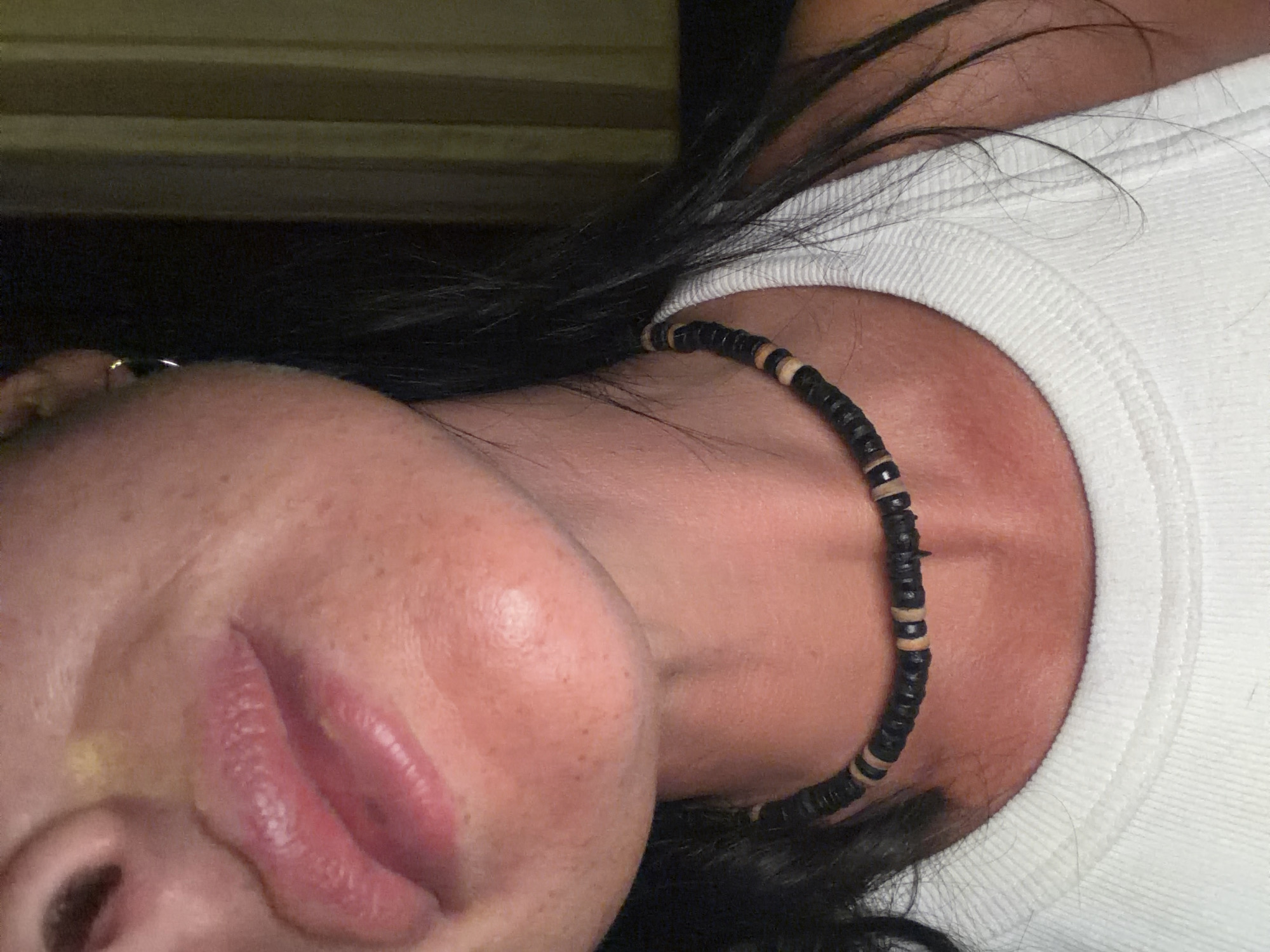 A close-up, tilted view focuses on a person's face and upper body. The person has fair skin, visible freckles, and full, pink lips. An earring is noticeable on the left ear. The person's black hair falls onto the right shoulder, which is covered by a white, crew-neck shirt. The person wears a black and beige beaded necklace around the neck. The background is dark. The location is unknown.