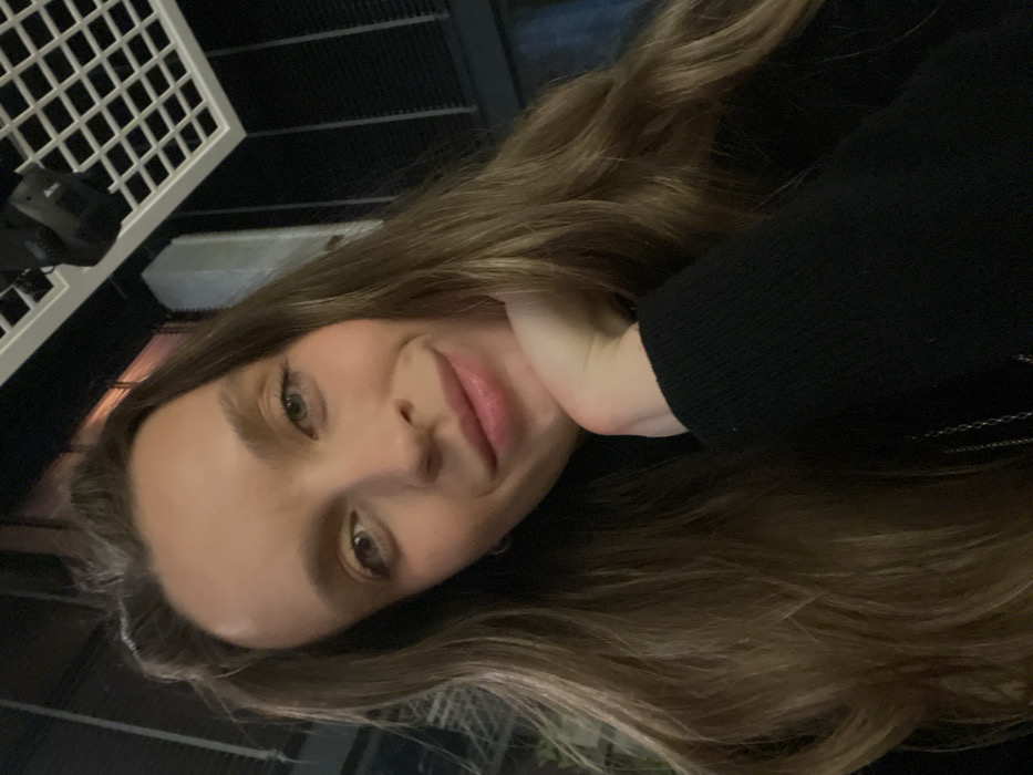 A woman with long brown hair and light skin poses for a selfie. She is wearing a black long-sleeved shirt. Her left hand is under her chin. The background is dark, and part of a white latticework structure is visible in the upper left corner. The image is slightly tilted to the right. She has light eyes and pink lips.