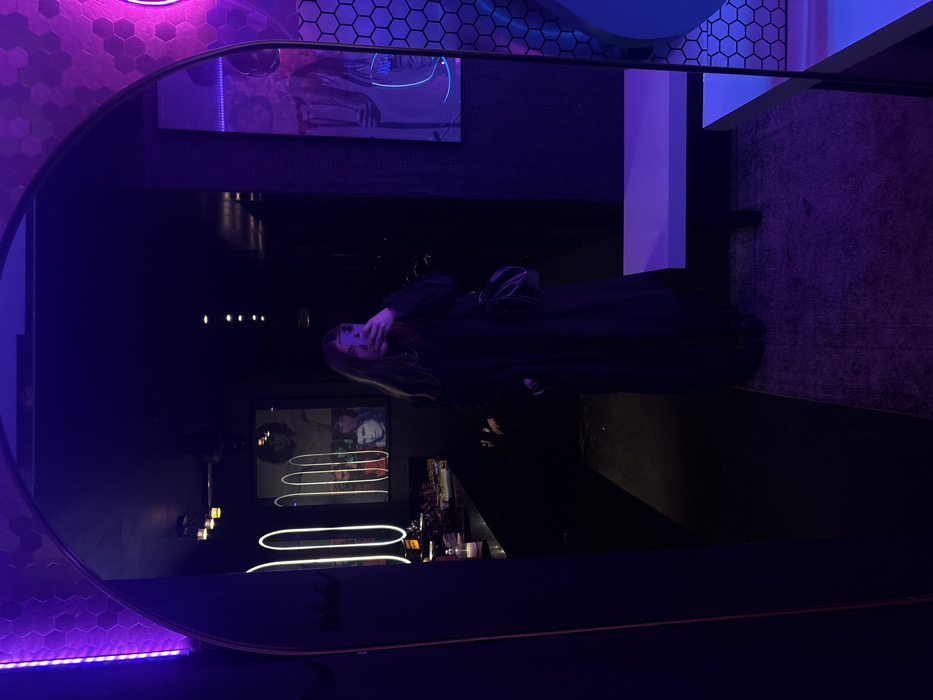 A long-haired individual is captured in a mirror selfie within the dimly lit interior of Bar El Bosque, located in the Roma Norte neighborhood of Mexico City, Mexico. The individual, dressed in a long, dark garment and carrying a dark shoulder bag, holds a smartphone with a light-colored case at chest height to photograph their reflection in a large, arched mirror. The environment is characterized by intense purple and pink neon lighting. To the left within the reflection, a counter is visible with several items, illuminated by vertical white neon light strips forming elongated oval shapes. Above the counter, a framed poster features multiple faces, including a prominent depiction resembling Jim Morrison. To the right, a wall section is covered in hexagonal tiles, further illuminated by a pink neon light. A white bench extends along the lower right side. The floor beneath the individual is covered by a patterned rug, transitioning to a darker surface in the foreground.