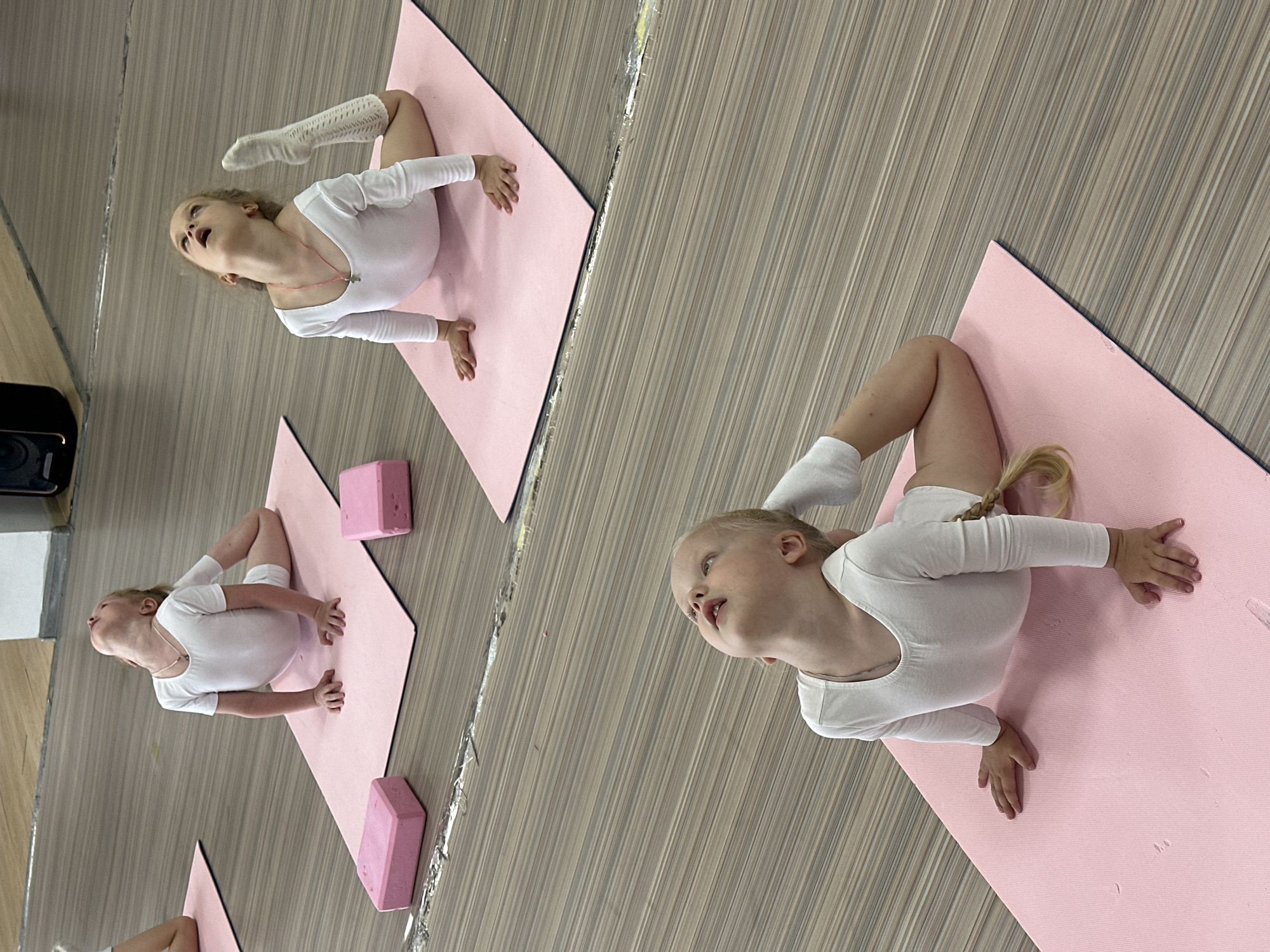 The image depicts three young girls in white leotards practicing gymnastics on pink mats in what appears to be a studio or dance hall with a wooden floor. The girl in the foreground is performing a backbend, with her hands on the mat and her head tilted back. The girl in the middle ground is also in a backbend, looking upwards, with one leg extended. The girl in the background is in a similar pose, looking upwards. The environment suggests a place for physical training or dance. There are pink yoga blocks on the mats. The lighting is bright and even, likely from overhead sources. The image's composition focuses on the girls and their poses, with the background slightly blurred. There are no discernible landmarks or clues to pinpoint the exact location within Votkinsk, Russia.