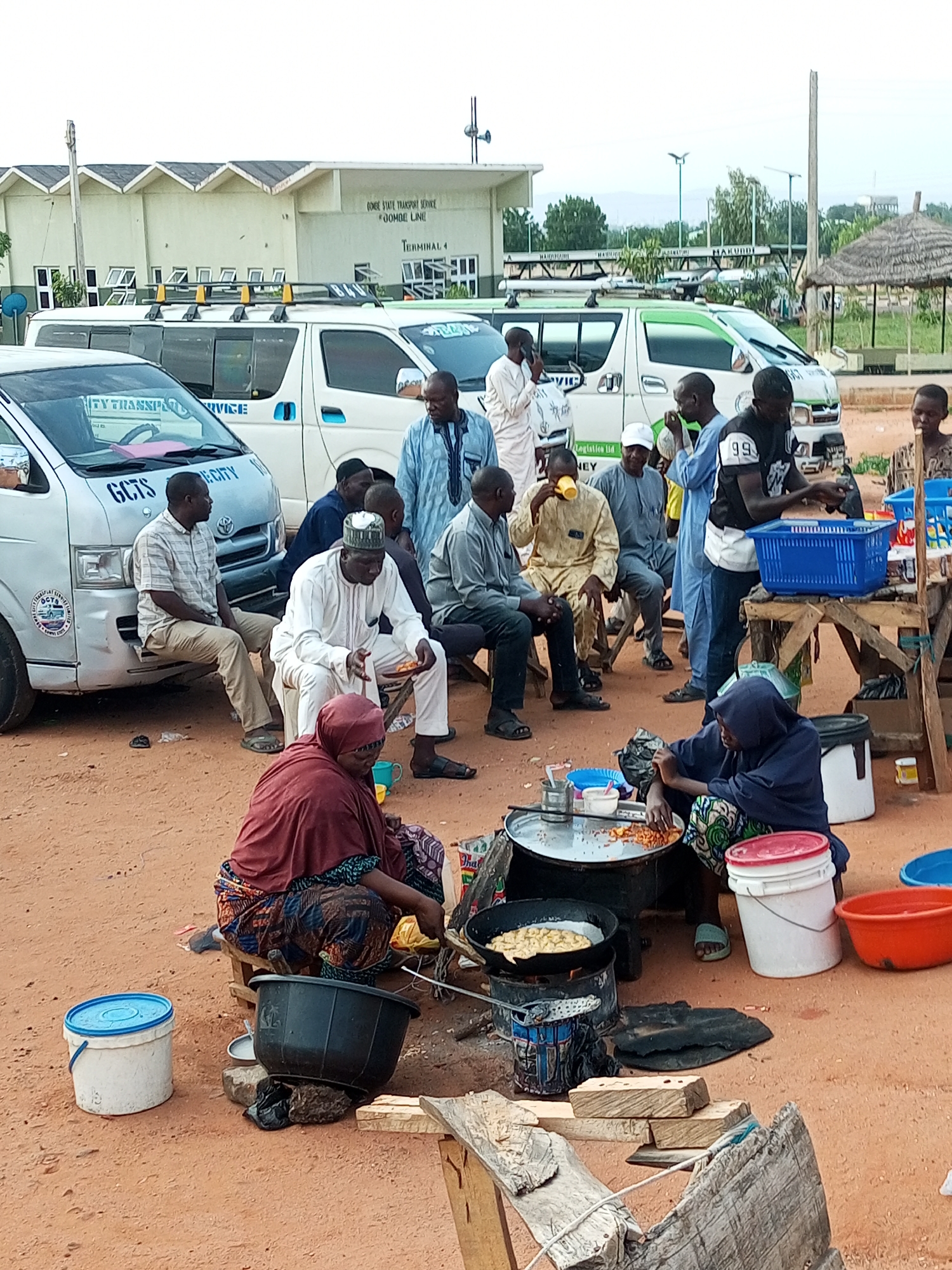 The image depicts an outdoor scene at Terminal 4, Gombe State Transport Service, Jomaoe Line in Gombe, Nigeria. Several white passenger vans are parked on a dirt ground. A group of men, some seated on low stools and others standing, are gathered around some food preparation. One man in a yellow garment is drinking from a yellow cup. Another man, wearing a white cap and traditional attire, is holding a plate with food. A woman in a dark blue headscarf is crouched near a makeshift cooking setup, preparing food on a large metal pan over a fire. Several buckets, including a white one with a blue lid and a black one, are visible. In the background, a long, light-colored building with a dark roof and a thatched-roof structure suggest the presence of amenities. The overall atmosphere appears to be a busy transportation hub with food vendors attending to travelers.
