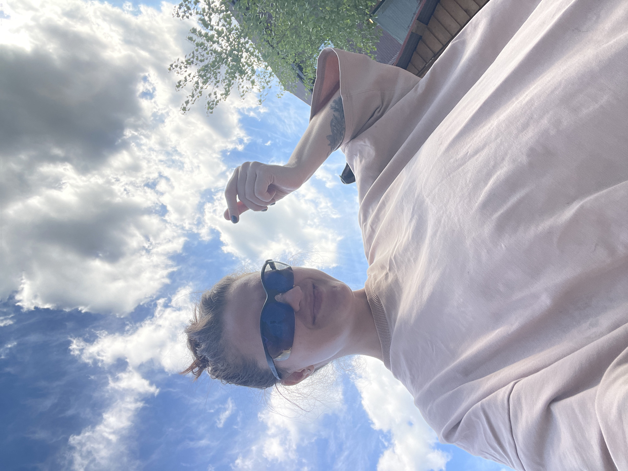 A low-angle shot of a woman wearing sunglasses and a light pink sweatshirt. She is standing outside, looking down at the camera with a sky full of clouds behind her. Her left arm is raised, and a tattoo is visible. A building and some trees are in the background. The image was taken in Misaylovo, Russia.