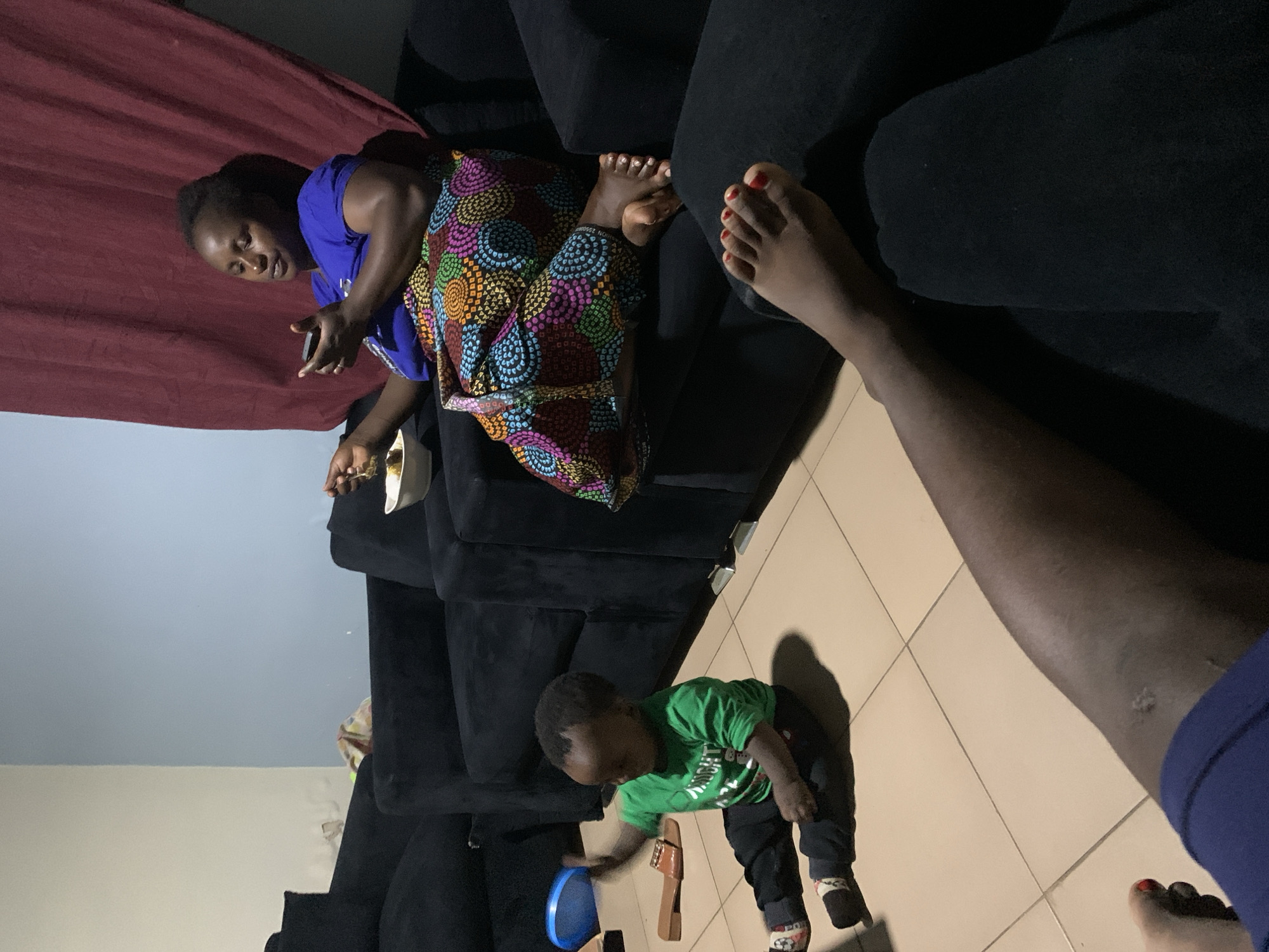 In an indoor setting, two individuals, an adult and a toddler, are present. The adult, a woman, is seated on a black couch and appears to be holding a phone and a bowl. She is wearing a blue shirt and a colorful skirt. A toddler, dressed in a green shirt and black pants, is sitting on the tiled floor. A blue object and a sandal are visible on the floor near the toddler. Another person's legs are visible in the foreground, resting on a couch. The environment includes a black couch, tiled flooring, and a dark red curtain in the background.