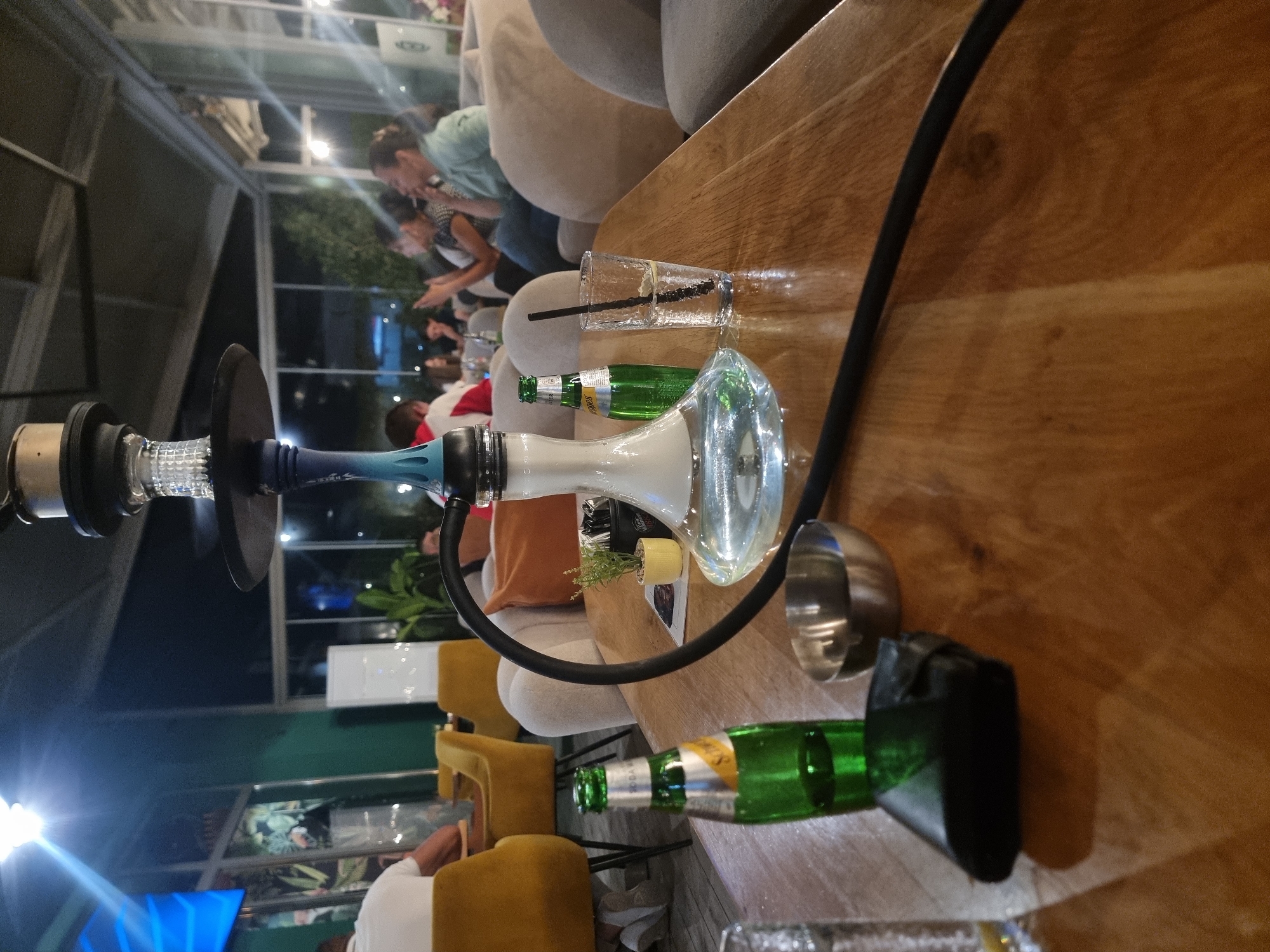 The image shows a hookah on a wooden table in an outdoor setting, possibly a cafe or lounge. In the background, people are seated, suggesting a social gathering. There are drinks on the table, including a glass with a straw and green bottles. The environment appears relaxed and open-air. Given the prompt's request for a location in Gotse Delchev, Bulgaria, it can be inferred that this scene is likely taking place in such a venue within that city. The lighting suggests it's either dusk or early evening. There are armchairs and decorative plants visible in the background, enhancing the ambiance. The camera angle is relatively close to the hookah and the drinks, emphasizing them as the focal points.