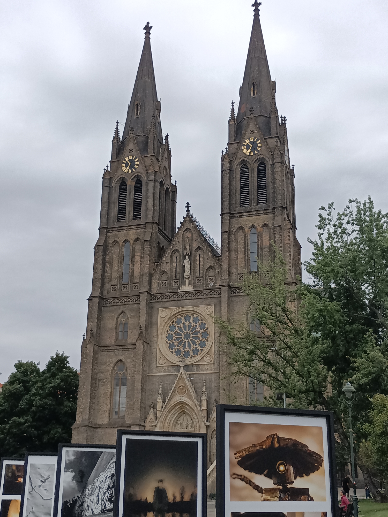 The image captures a view of the Basilica of Saint Peter and Saint Paul, located in Vyšehrad, Prague, Czechia. The church features two prominent spires, each topped with a cross. A large clock face is visible on the left spire, and another on the right spire. In the foreground, a series of framed photographs are displayed outdoors. The sky is overcast and grey. Lush green trees are present on the right side of the image, partially obscuring the church. A lamppost stands near the trees. Two individuals, one wearing a pink shirt and the other a dark top, are visible in the background on the right side.
