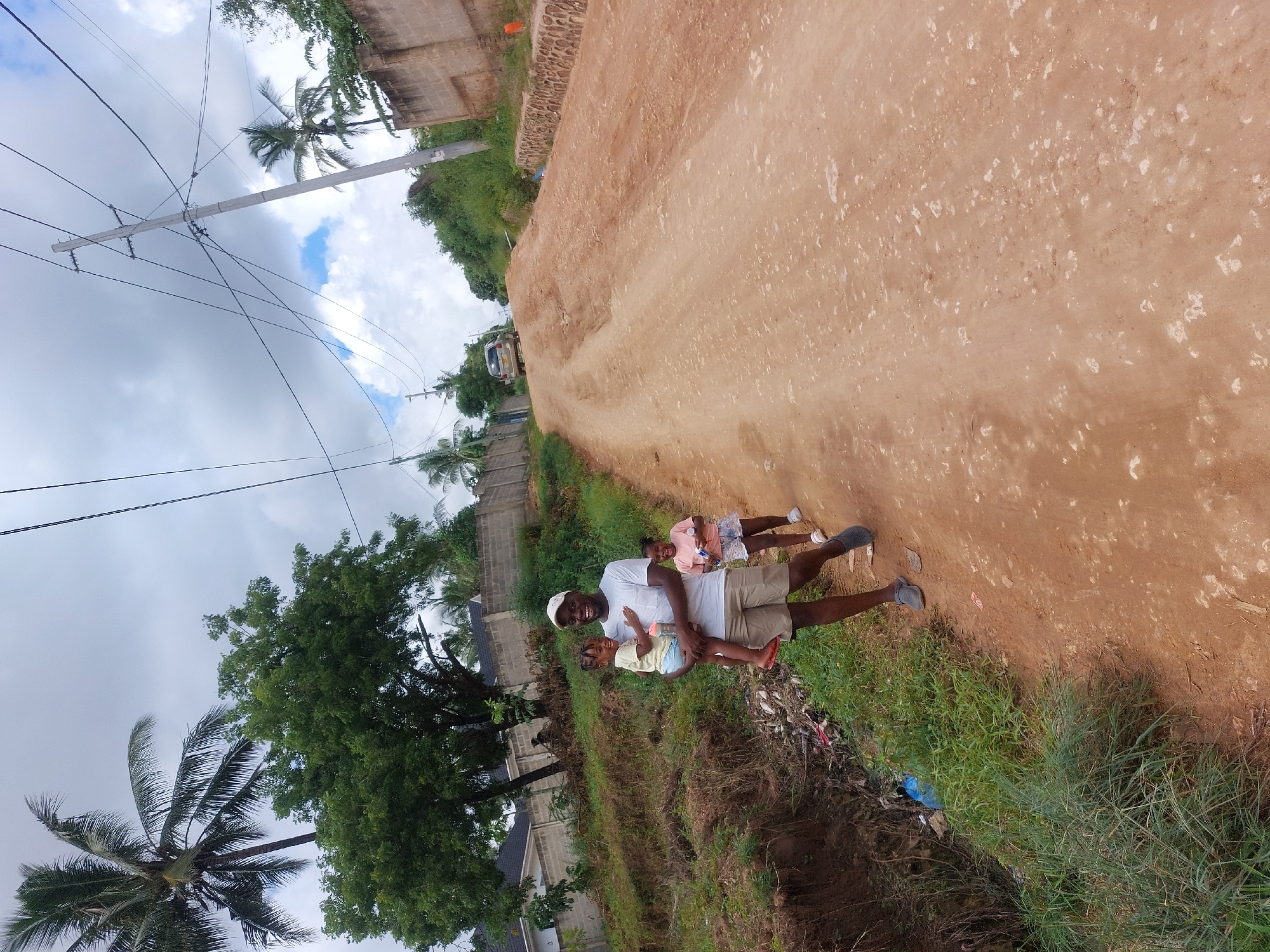 The image shows a man holding two young children on a dirt road next to a grassy embankment. The man is wearing a white t-shirt, khaki shorts, and a white baseball cap. The children appear to be female and are wearing light-colored clothing. They are all smiling. The road is unpaved and reddish-brown. To the right of the road, a grassy embankment slopes down to what appears to be a small ditch. Trash and debris are visible in the ditch. Behind the group, there are trees and buildings. In the background, a car is parked on the road. A palm tree and power lines are visible in the upper left of the frame.
While I lack specific details to definitively pinpoint the exact location in Dar es-Salaam, Tanzania, the environment suggests a residential area on the outskirts of the city, where unpaved roads and less developed infrastructure might be common. The presence of palm trees indicates a coastal region.