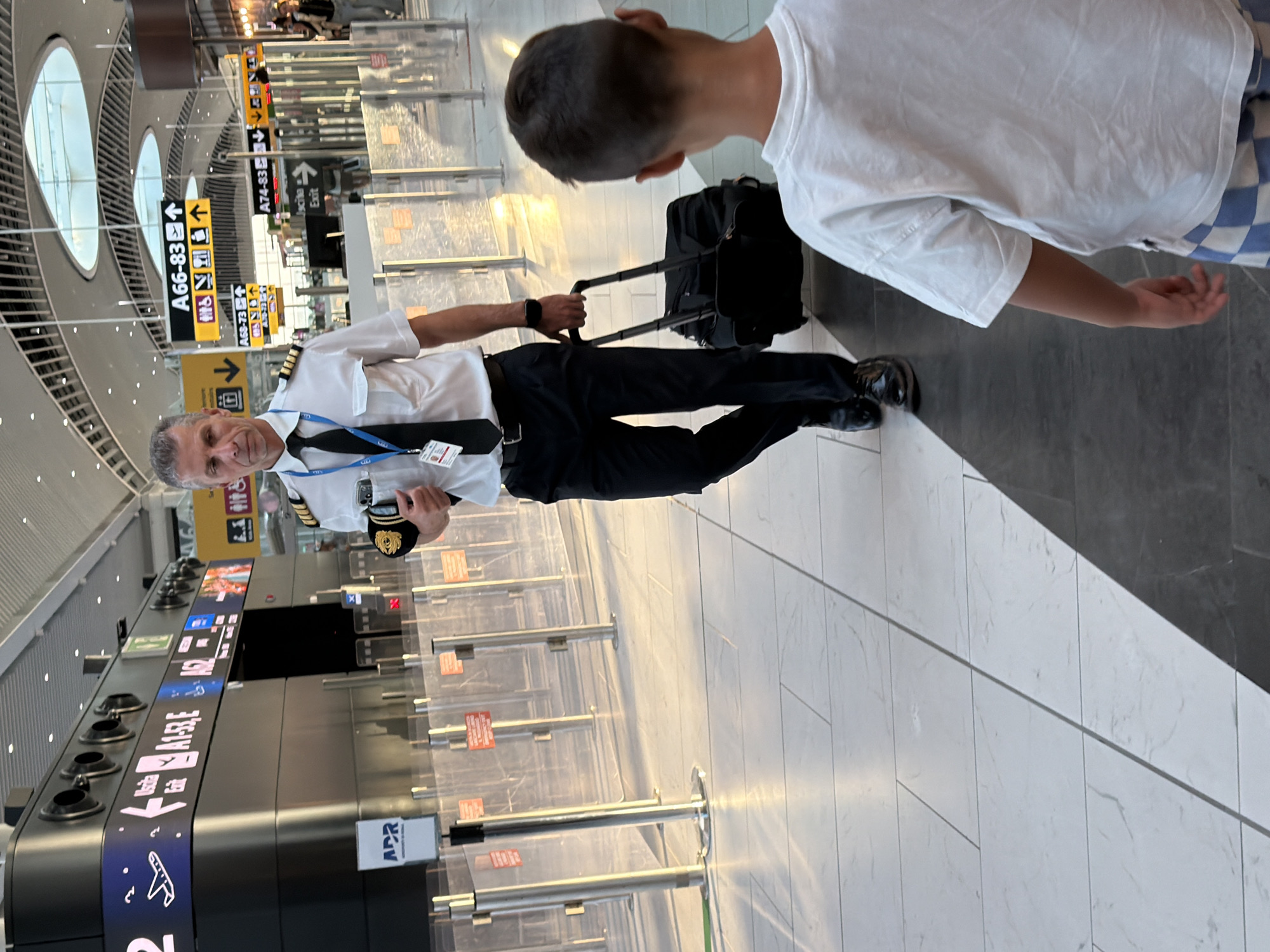 In Fiumicino, Italy, within a modern airport terminal, a male pilot in uniform, distinguished by epaulets on his shoulders and a black tie, walks with a rolling suitcase. He is captured mid-stride, his gaze directed forward. A young boy, facing away from the camera, stands in the foreground, his back to the pilot, wearing a white t-shirt and checkered shorts. The airport environment is characterized by sleek architecture, illuminated signage indicating flight gates such as "A66-83" and "A74-83," and a polished, tiled floor. Sunlight streams into the terminal, casting long shadows and creating a bright, airy atmosphere. Transparent barriers and stanchions are visible, suggesting a secure waiting or boarding area.