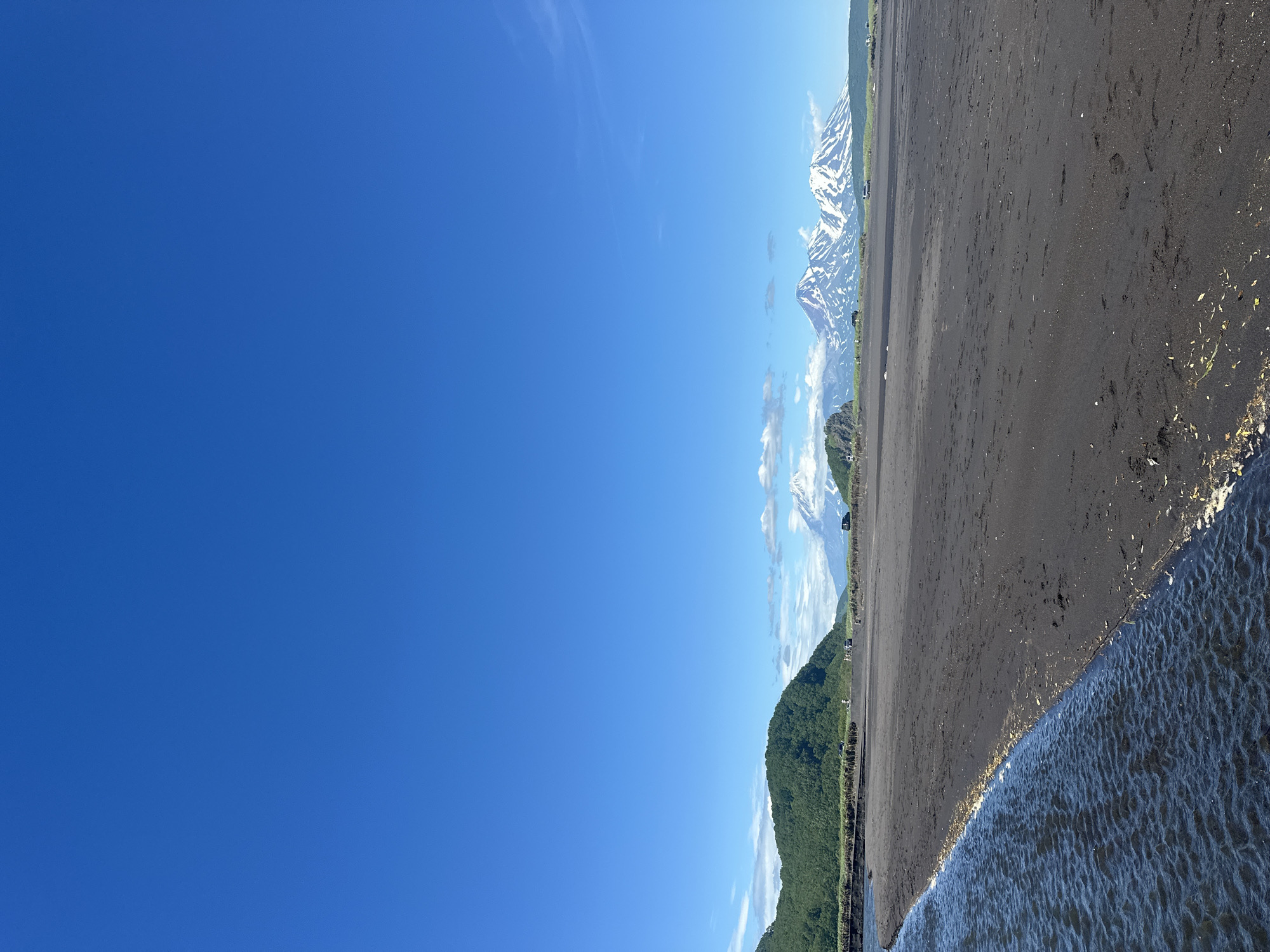 The image depicts a wide expanse of black sand beach leading to the ocean, with a clear blue sky overhead. In the distance, a range of snow-capped mountains is visible. There are several vehicles and a few people scattered along the coastline. The scene appears to be a natural landscape near Petropavlovsk-Kamchatsky, Russia, likely a popular tourist spot. The black sand suggests volcanic activity in the region, a characteristic feature of Kamchatka. The water in the foreground is dark and choppy, contrasting with the calm sky and the majestic mountains. The overall impression is one of natural beauty and rugged wilderness.