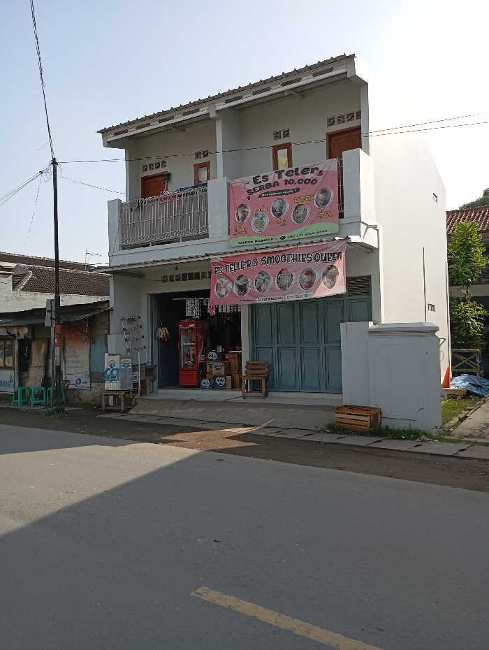 The image depicts a two-story, light-colored commercial building, located in Cipatik, Indonesia, viewed from a street perspective under a clear, bright sky. The building's facade is dominated by two large banners: an upper red banner advertising "Es Teler SERBA 10.000!" and a lower pink/purple banner reading "ES TELLER & SMOOTHIES QUEEN," both featuring images of various beverages. The ground floor shop entrance on the left is open, revealing a red commercial refrigerator, stacked boxes, and several small green plastic stools. The right side of the ground floor is secured by closed blue bifold doors. The second floor features a balcony with a metal railing, accessed by two brown doors. To the left of the main building, a portion of an older, single-story building with a dark tiled roof is visible. The foreground consists of an asphalt road with faint yellow markings and a concrete sidewalk. Strong sunlight casts clear shadows across the road and sidewalk. No individuals are present in the scene.