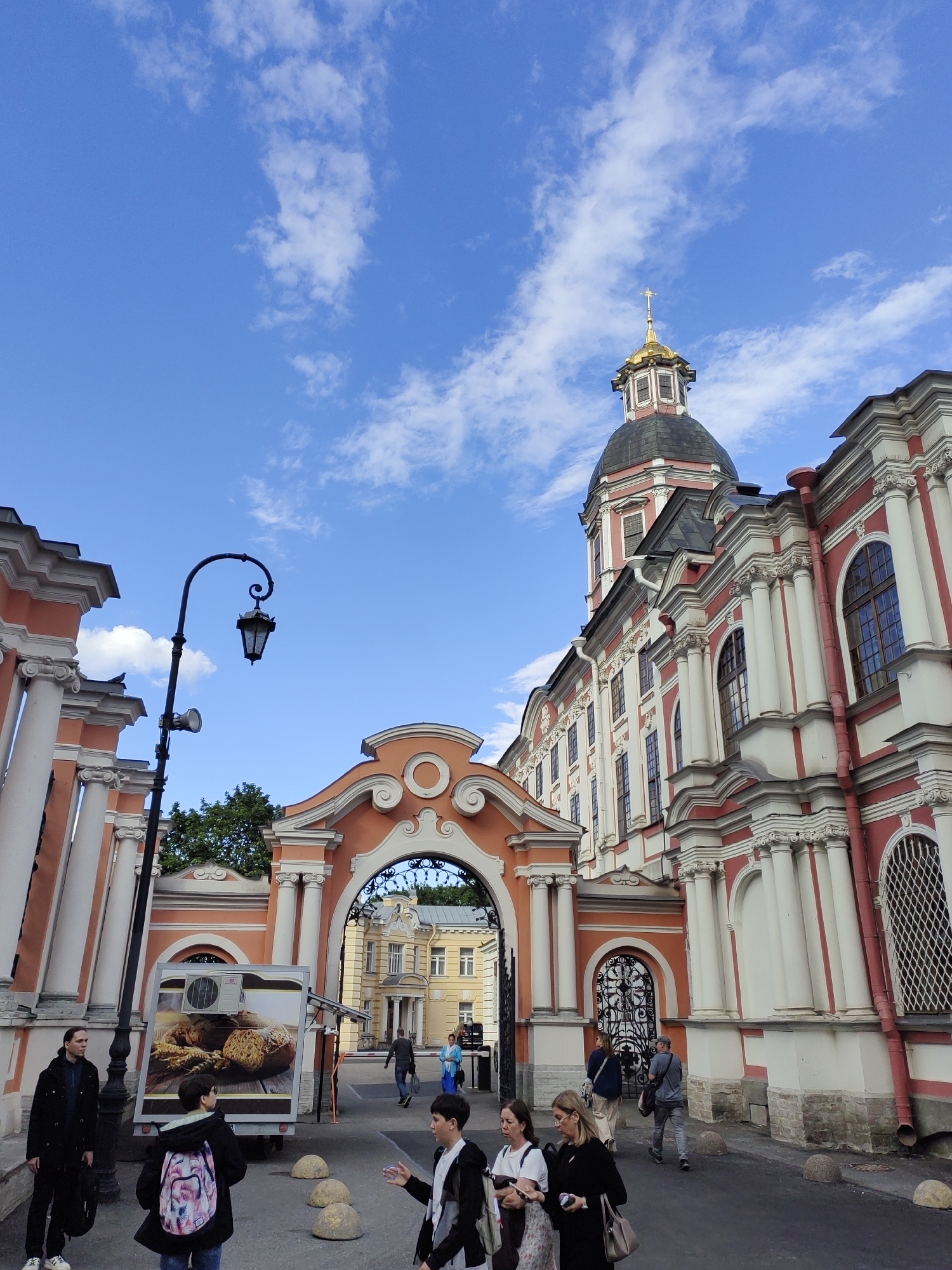 This image depicts a street scene in front of a grand, ornate building, likely a historical church or monastery, under a bright blue sky with scattered white clouds. The architecture is predominantly white and red, featuring classical columns, arched windows, and a prominent golden dome topped with a cross. An arched gateway with decorative ironwork leads to the building.
Several people are visible in the foreground and midground. On the left, a person in a black coat stands near a lamp post. In the center, a group of younger individuals are walking and interacting, one with a backpack adorned with a colorful pattern. Further back, more people are seen walking, including a person in a blue headscarf.
To the left of the entrance, a food truck with an advertisement for bread is parked. The overall atmosphere appears to be that of a typical day in a tourist or public area, with people moving about. The clear sky suggests it is daytime, likely late morning or afternoon, with pleasant weather. The architectural style and the presence of a religious building strongly suggest the location is in Russia, specifically Saint Petersburg, as indicated by the context. No discernible text is visible in the image.