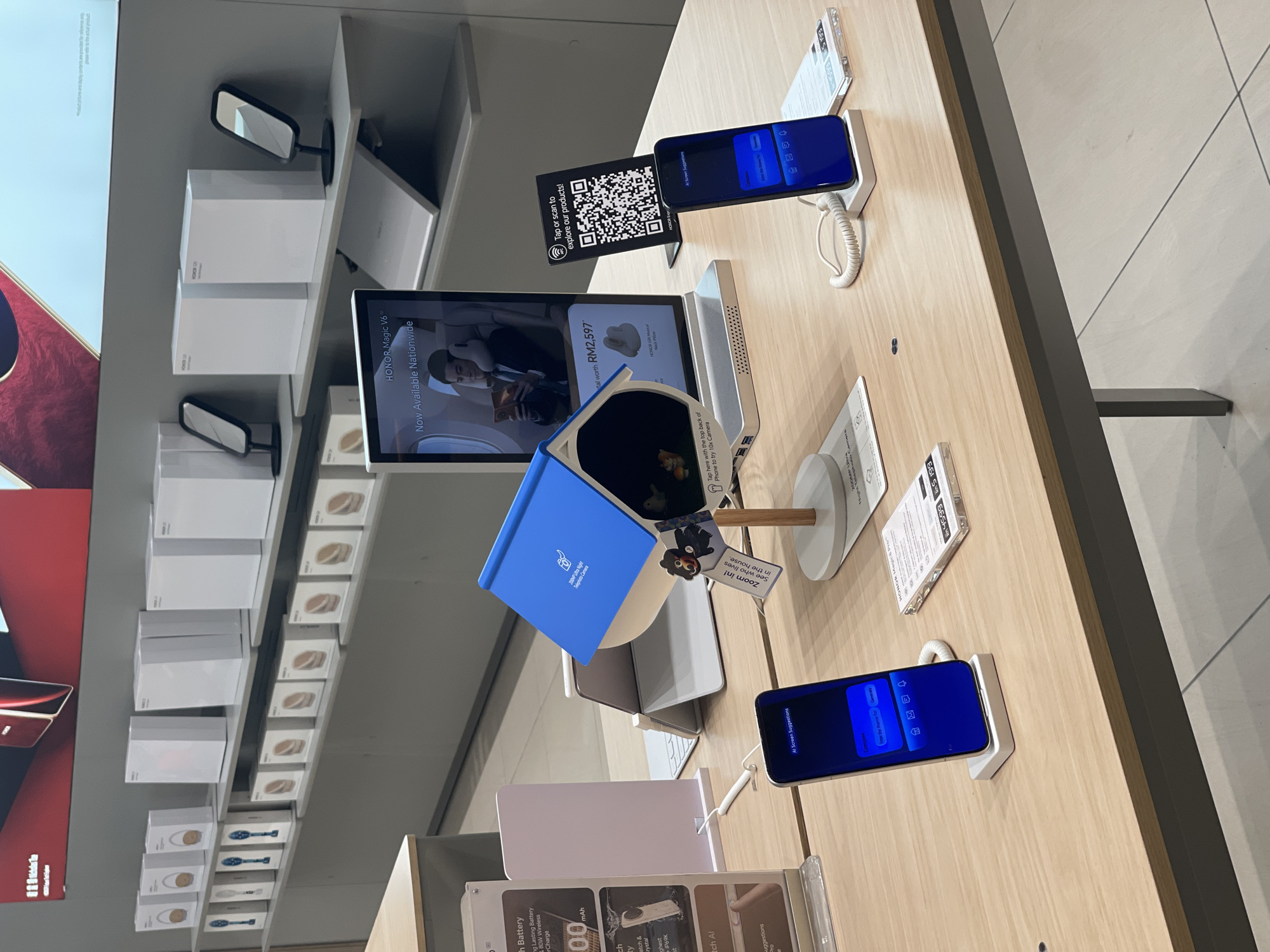 The image shows a retail store in Kuala Lumpur, Malaysia, displaying mobile phones and accessories. A dark blue HONOR smartphone is prominently featured on a white display stand in the foreground, with its screen showing "AI Screen Suggestions" and options for "Companion," "Enter the Theme text," and "Generate." Another similar smartphone is located to the right of the first, also on a white display stand, displaying a QR code for product exploration. Behind the smartphones, a blue and white birdhouse-like structure serves as a display for small figurines. Further back, a tablet or larger display screen advertises "HONOR Magic V6" and states "Now Available Nationwide" with a price of RM2,597. Shelves in the background are stocked with white boxes, some with the HONOR branding visible, and display stands holding mirrors. The overall environment suggests a modern electronics store.