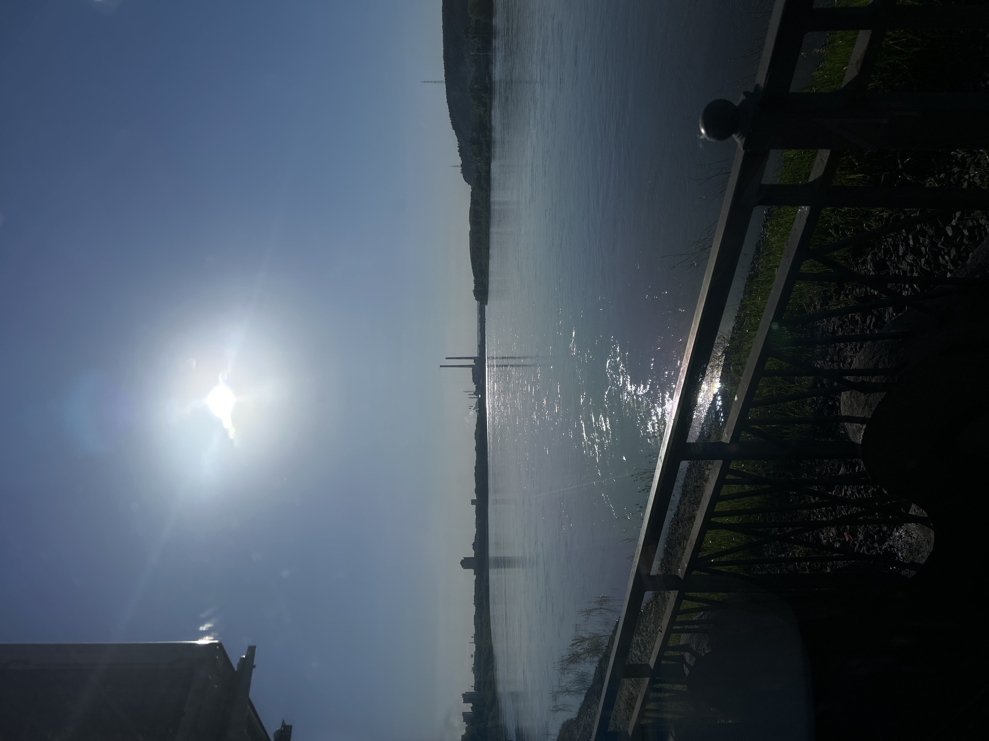 The image, originally rotated 90 degrees clockwise, depicts a wide, sunlit outdoor scene featuring a body of water, likely a river or large lake, viewed from a bank or promenade. In the foreground, a dark metal railing runs diagonally across the lower right of the frame, with glimpses of shadowy ground cover, including grass and small stones, visible beneath it.
The sun is bright and prominent in the upper left portion of the sky, casting a strong glare and producing visible lens flares. Its intense light reflects brilliantly off the water's surface, creating a shimmering, sparkling effect that stretches across the calm water.
On the distant horizon, a long, low line of dark green hills or dense treeline is visible. Along this far bank, several indistinct structures can be observed, including what appear to be two tall, thin industrial smokestacks or pillars in the mid-distance. Further along the horizon, faint silhouettes of other buildings suggest a distant urban or industrial presence. The sky above is a clear, light blue, indicating bright, sunny weather. The time of day appears to be midday or early afternoon due to the sun's high position and intensity. No people are visible, and there is no visible text. The scene conveys a sense of openness and natural expanse under a bright sky.