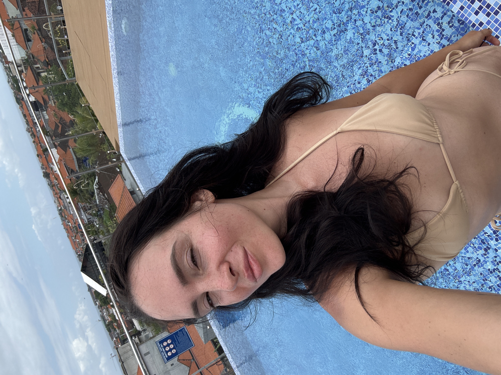 A woman with long, dark hair and freckles is taking a selfie next to a rooftop swimming pool. She is wearing a light brown bikini. In the background, there is a city skyline with red-tiled roofs and lush green trees. The sky is cloudy. A sign with rules for the pool is visible on the left side of the image. The pool water is blue and tiled.