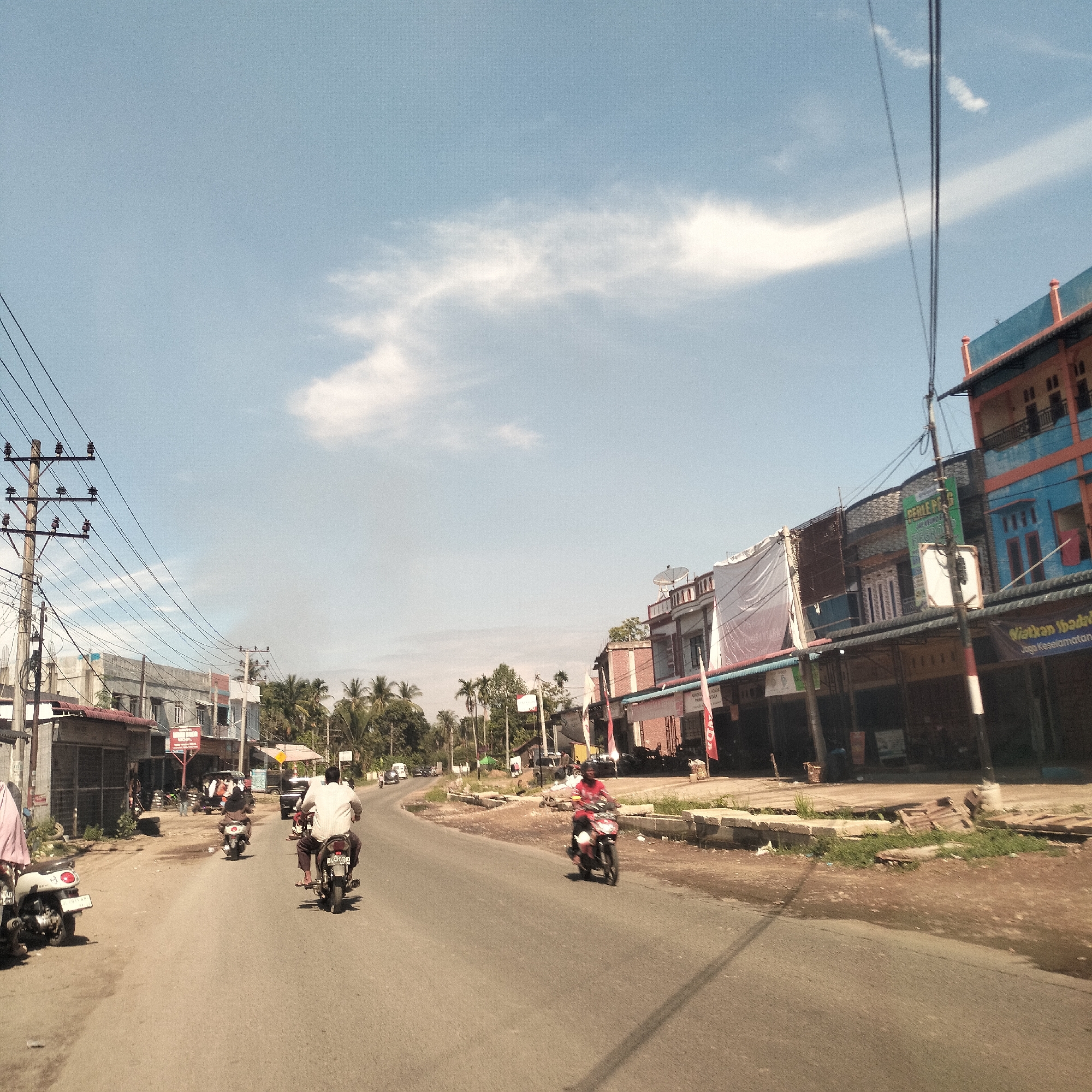 This image captures a bright, sunny day on a busy street in Manetunong, Indonesia. The scene is dominated by a paved road that curves slightly to the right, with buildings and vegetation lining both sides.
Several people are visible, primarily riding motorcycles or scooters, indicating daily commute or travel. On the left side of the road, a person on a white scooter is partially visible in the foreground, with another rider on a darker motorcycle further down. In the middle-ground, a person in a white shirt rides a motorcycle away from the camera, with a partially visible license plate starting "BK 4". On the right, a person in a red shirt rides a motorcycle, approaching the viewer.
The street is lined with a mix of single and multi-story buildings, many appearing to be commercial shop-houses with various architectural styles and colors, including blue, orange, and grey. Some buildings feature awnings or banners, and white tarpaulins are draped over parts of others. Overhead, numerous power lines crisscross the blue sky, supported by utility poles that are prominent on both sides of the road. Palm trees and other tropical foliage are abundant in the background, especially on the left.
Visible text includes a green sign on a pole on the right reading "PERLE PR..." and "SALON KECANTIKAN" (Beauty Salon). Another banner on a blue and orange building to the right shows "NIATKAN IBADAH" and "Jaga Keselamatan". A red flag with a logo resembling "IDA" is also visible. The clear sky with scattered white clouds, combined with the bright lighting, suggests it is mid-morning or early afternoon.
