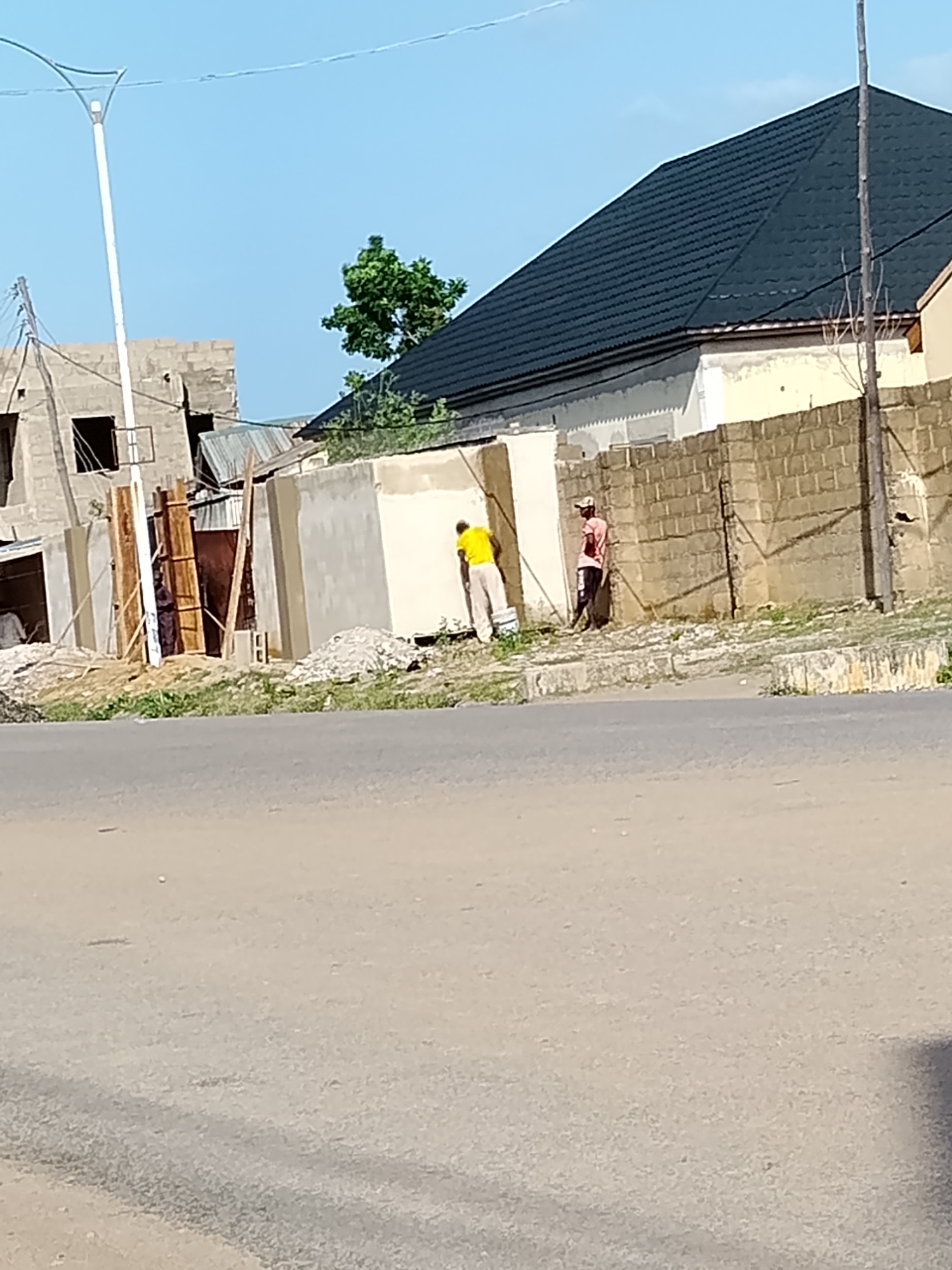 In Wauro Leggal, Nigeria, two men are seen by a construction site. The man on the left, wearing a yellow shirt and white pants, is painting a light-colored wall. To his right, a man in a pink shirt and dark pants is standing near a brick wall. The construction site includes a partially built structure with cinder blocks, wooden supports, and corrugated metal roofing. A dark tiled roof tops a nearby finished building. The scene is set against a clear blue sky, with a street in the foreground and sparse vegetation.