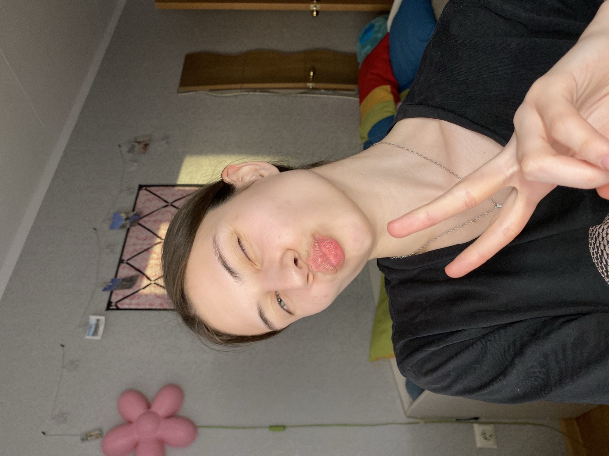 A young person with fair skin, dark hair, wearing a silver chain, and a black top is making a peace sign with their hand and a kissing face towards the camera. They are indoors. A pink flower decoration is on the wall, as well as a grid-like decoration. The person is likely in a bedroom. The city of Votkinsk, Russia, is not apparent from the given information.