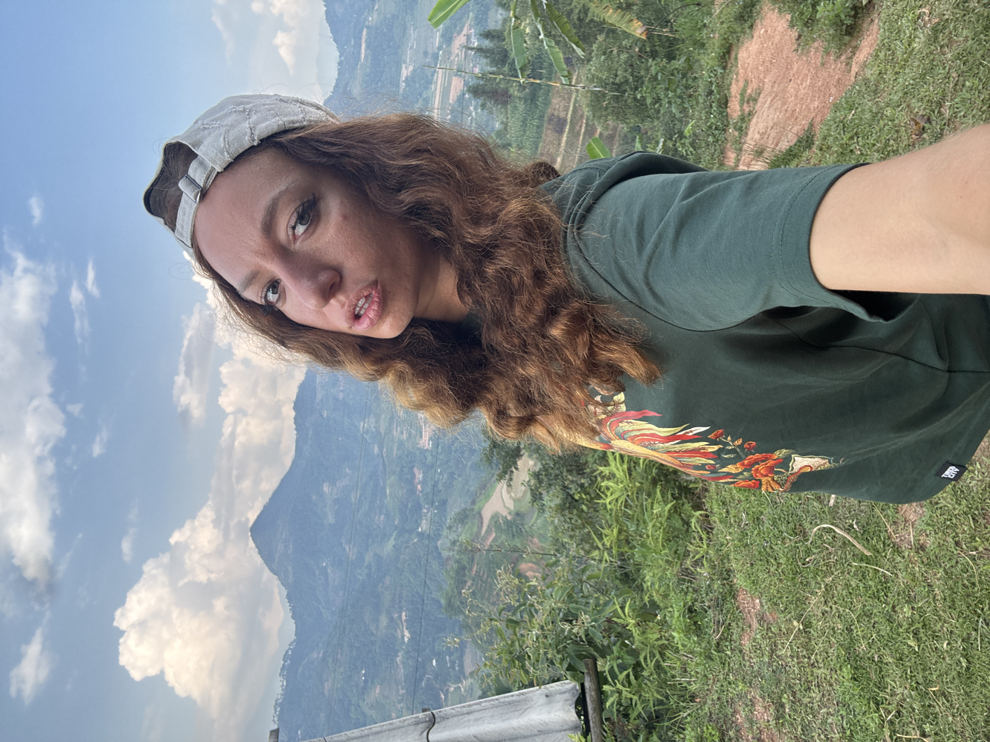 A woman with curly brown hair and a light-colored baseball cap is taking a selfie. She is wearing a dark green t-shirt with a colorful design on it. The background shows a mountainous landscape under a partly cloudy sky. The scene appears to be in Pờ Ly Ngài Commune, Vietnam, suggested by the vegetation and terrain visible in the background. Some cultivated fields and possibly rice paddies are discernible in the distant landscape. There is also a metal railing on the left side of the image, suggesting she might be near a viewpoint or road. The ground is covered with green grass and other vegetation.