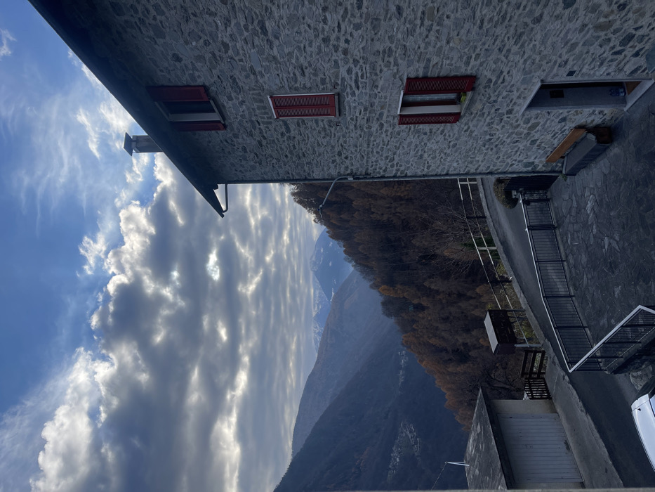 The image, captured in Dosso (Ciappanico), Italy, presents a scenic view of a mountainous landscape from an elevated perspective. A prominent stone-built structure, likely a house or chalet, occupies the left side of the frame. Its grey, rough-hewn stone facade features multiple windows, each adorned with vibrant red louvered shutters, mostly closed. A chimney stack is visible on the roofline.

The sky, filling the upper right portion of the scene, exhibits a striking combination of clear blue patches and expansive white to grey stratocumulus clouds, with sunlight intermittently piercing through them.

In the mid-ground, a steep mountainside is densely covered with deciduous trees, displaying the muted brown and ochre tones characteristic of late autumn or early winter foliage. Beyond this, a series of imposing mountain ranges recedes into the distance, with the highest peaks exhibiting traces of snow.

On the immediate right, a paved area, possibly a narrow road or a stone-tiled courtyard, is visible, bordered by dark metal railings. Further down, what appears to be another building or a garage door can be seen.

The scene is devoid of people or any specific activity, conveying a tranquil and serene atmosphere. The time of day appears to be daylight, with moderate cloud cover but good visibility across the vast landscape. The architectural style and natural elements strongly suggest a traditional alpine or pre-alpine setting. No text is visible in the image.
