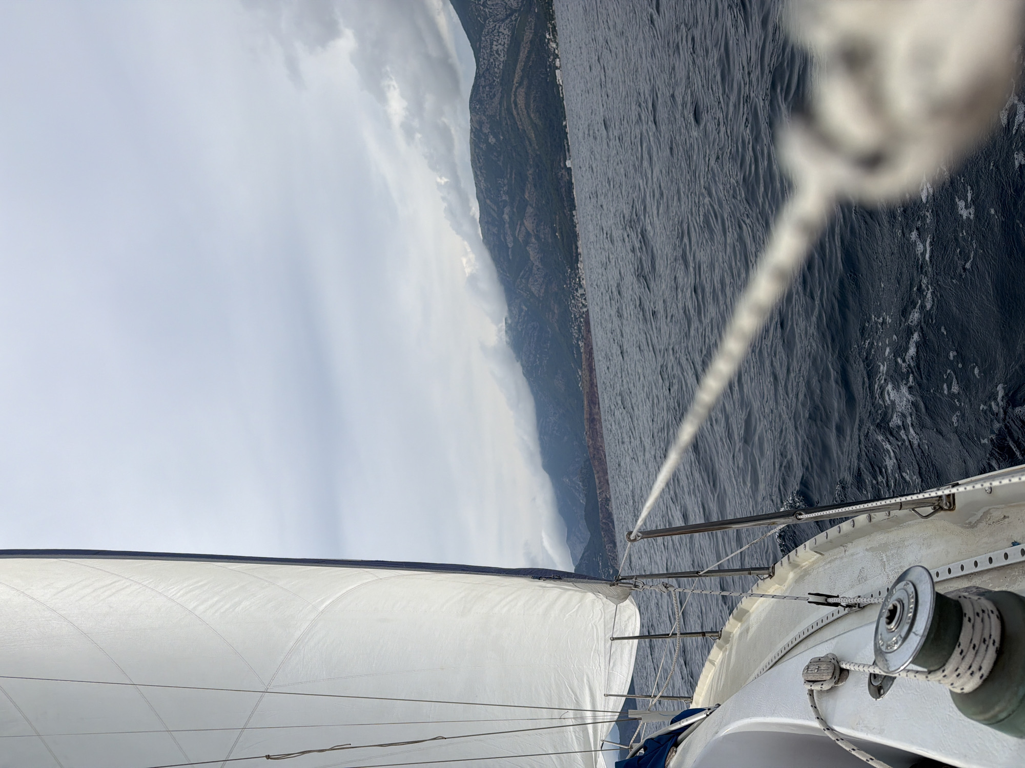A view from a sailboat sailing on dark blue water, with a large white sail dominating the left side of the frame. A thick rope extends diagonally across the foreground, leading towards the blurred front of the boat. In the background, a coastline with green, mountainous terrain is visible under a cloudy, overcast sky. Buildings are scattered along the base of the mountains. The scene appears to be the coast of Bar, Montenegro.
