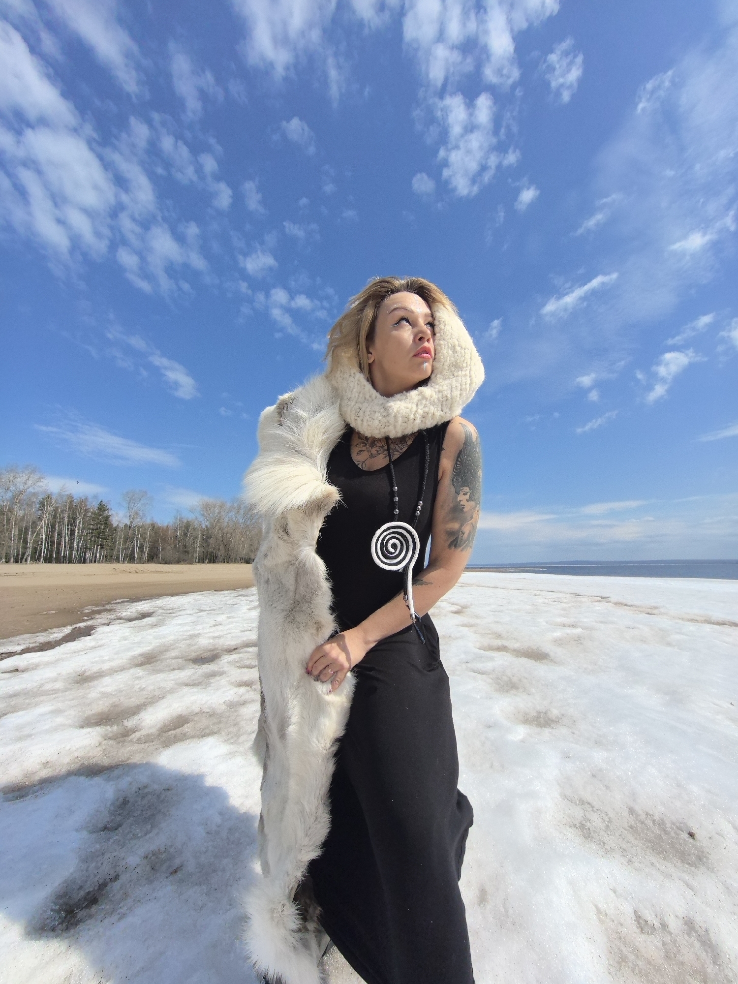 A medium shot captures a woman standing outdoors in what appears to be a melting, snowy landscape, likely near a body of water in Noviy, Russia. She has blonde hair and is looking upwards and to her right, with a contemplative expression. She wears a black sleeveless dress or tank top, a thick, white or cream-colored chunky knit cowl around her neck, and a long, white, furry stole draped over her left arm and shoulder. Visible tattoos adorn her arms, and she has a lip piercing. A black beaded necklace with a prominent white spiral pendant hangs from her neck.
The ground around her is covered in patches of snow and ice interspersed with areas of brown soil or sand. In the background to her left, a bare forest of tall, slender deciduous trees lines a bank. To her right, a vast expanse of water, possibly a sea or a large lake, stretches to the horizon, with some ice or snow visible along the shoreline. The sky above is a brilliant blue with scattered white, wispy clouds, indicating clear, sunny weather. The bright sunlight suggests it is daytime. No discernible text is visible in the image.