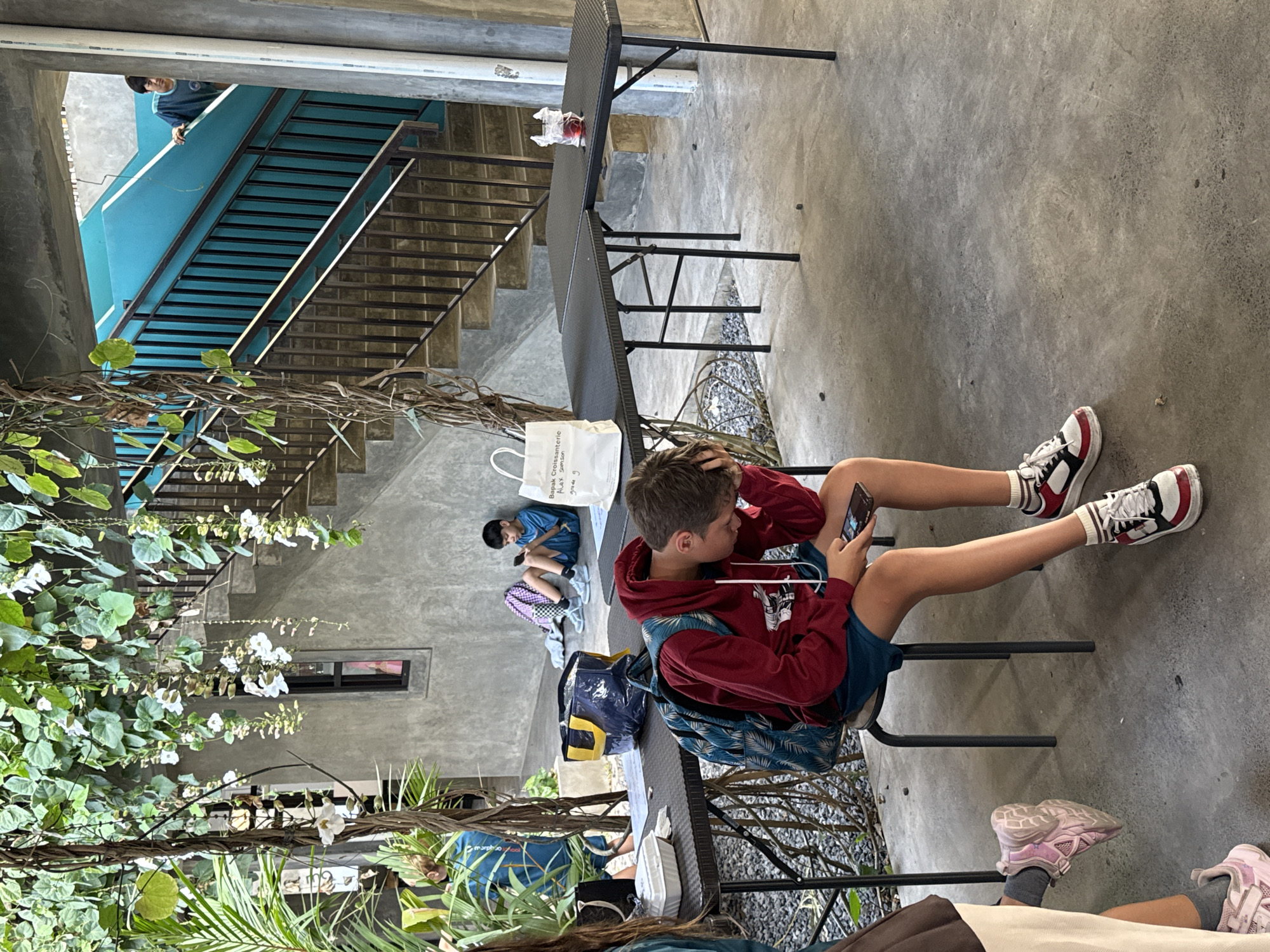 In North Kuta, Indonesia, within an outdoor courtyard, a young boy with brown hair is seated on a dark chair, engrossed in his mobile phone. He wears a red hooded sweatshirt, blue shorts, a backpack with a tropical pattern, and white and black sneakers with red accents. To his left, another boy in a blue t-shirt and shorts is sitting on the floor, also looking at a phone. Behind them, a purple and white backpack rests on the ground. In the background, a concrete staircase with dark metal railings ascends, painted a vibrant teal on the wall. Various tables are set up in the courtyard, some holding a white paper bag, a yellow and blue bag, and a clear plastic cup with a red substance. Green vines with white flowers hang from above, creating a natural canopy.