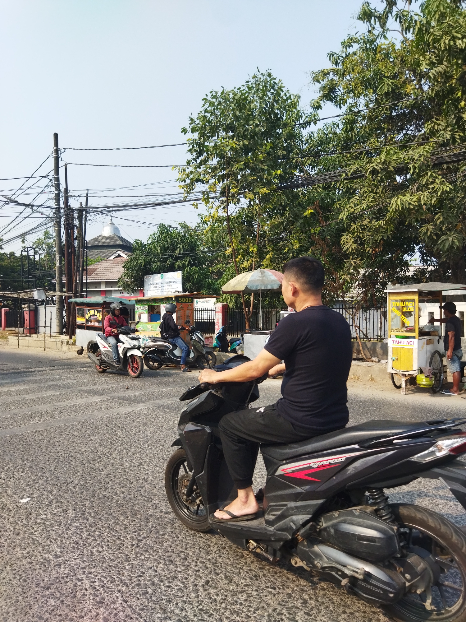 The image depicts a street scene in Pisangan Jaya, Indonesia, characterized by a clear, bright sky and lush green trees lining the road. In the foreground, a man wearing a black t-shirt and black pants rides a black motorcycle. He is wearing flip-flops. The motorcycle has a red and silver "VARIO" logo on its side. Behind him, two other individuals on motorcycles are visible: one in a red shirt and the other in a black hoodie, both wearing helmets. To the right, a food cart with a yellow and white structure is set up, displaying prices and menu items. A person in a dark shirt and shorts stands behind the cart, appearing to be serving food. Further in the background, a building with a domed, silver roof is partially obscured by trees, possibly a mosque. Utility poles with numerous wires are a prominent feature of the urban landscape, crisscrossing the sky. The road surface is paved with a textured, grey material.